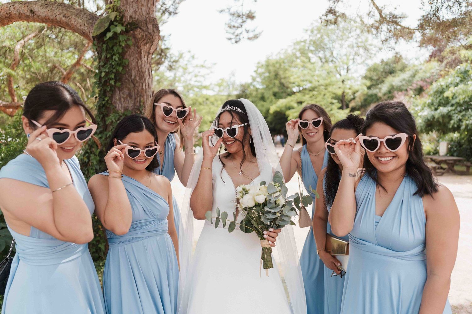 La mariée et ses demoiselles d'honneur en robes bleues, portant des lunettes de soleil en forme de cœur et tenant leur bouquet.
