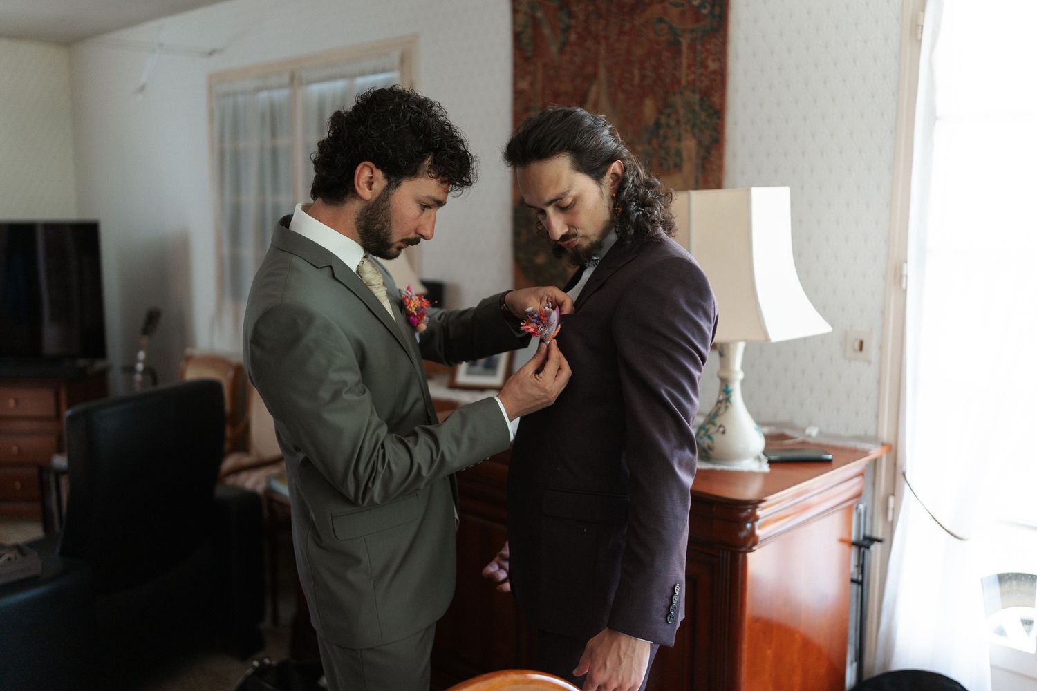 Two men in suits helping each other get dressed, one pinning a boutonniere on the other's lapel indoors.