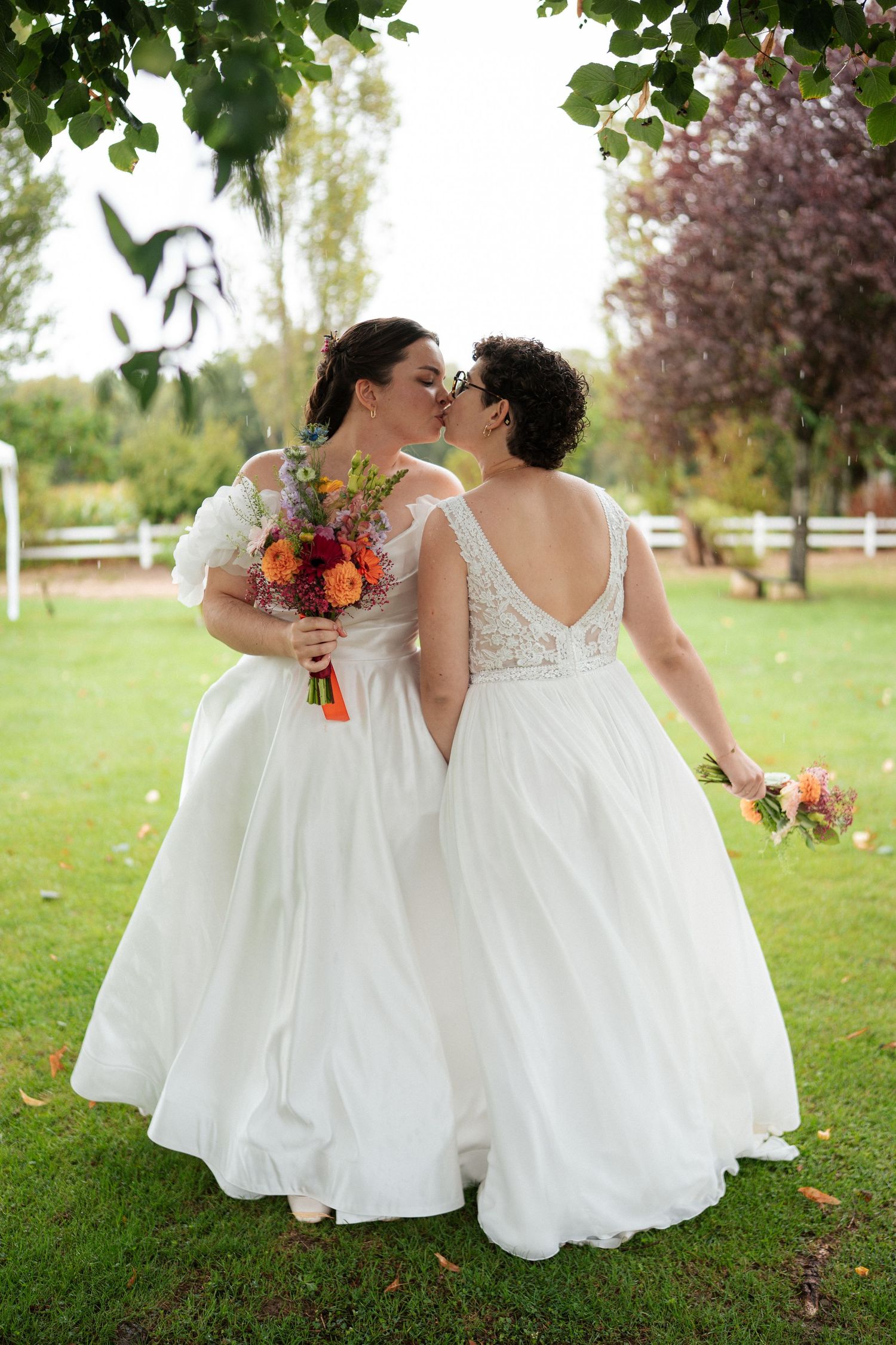 Two brides in white gowns share a kiss outdoors, holding colorful bouquets on their wedding day.