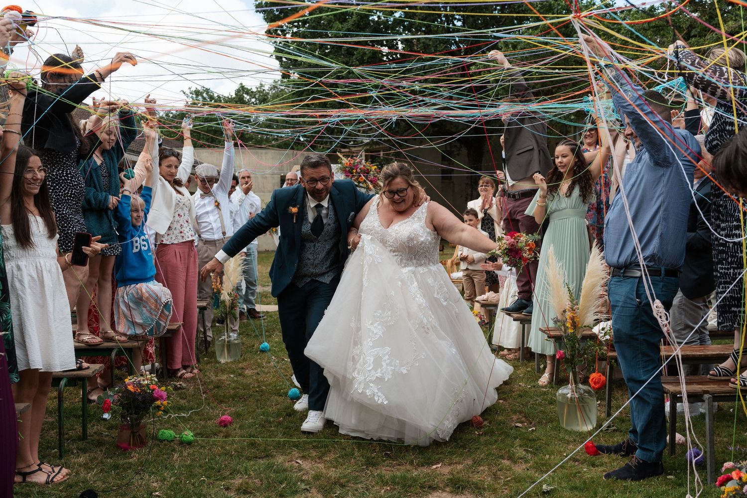 Joyful newlyweds run through colorful streamer tunnel surrounded by cheering guests at outdoor wedding celebration.