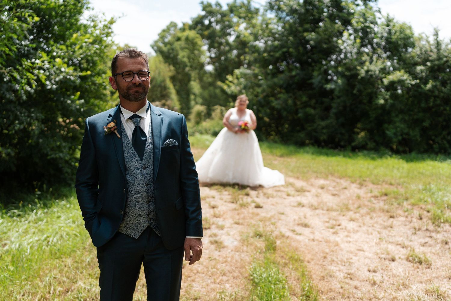 Groom in blue suit stands in field as bride in white gown approaches on sunny wedding day.