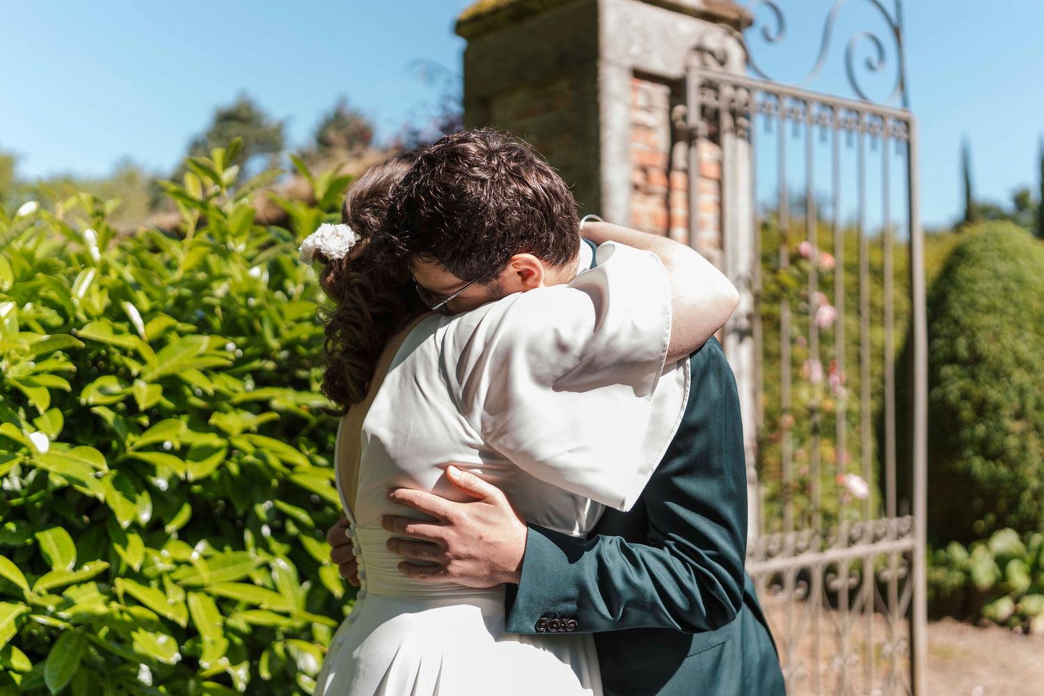 Un couple s'enlace tendrement près d'un portail en fer forgé orné, entouré d'une végétation luxuriante, par une journée de mariage