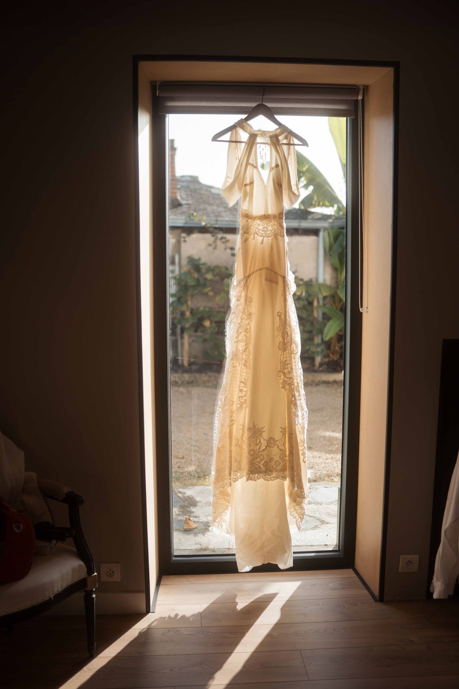 Élégante robe de mariée en dentelle suspendue à une fenêtre ensoleillée, laissant filtrer une lumière dorée à travers la vitre.