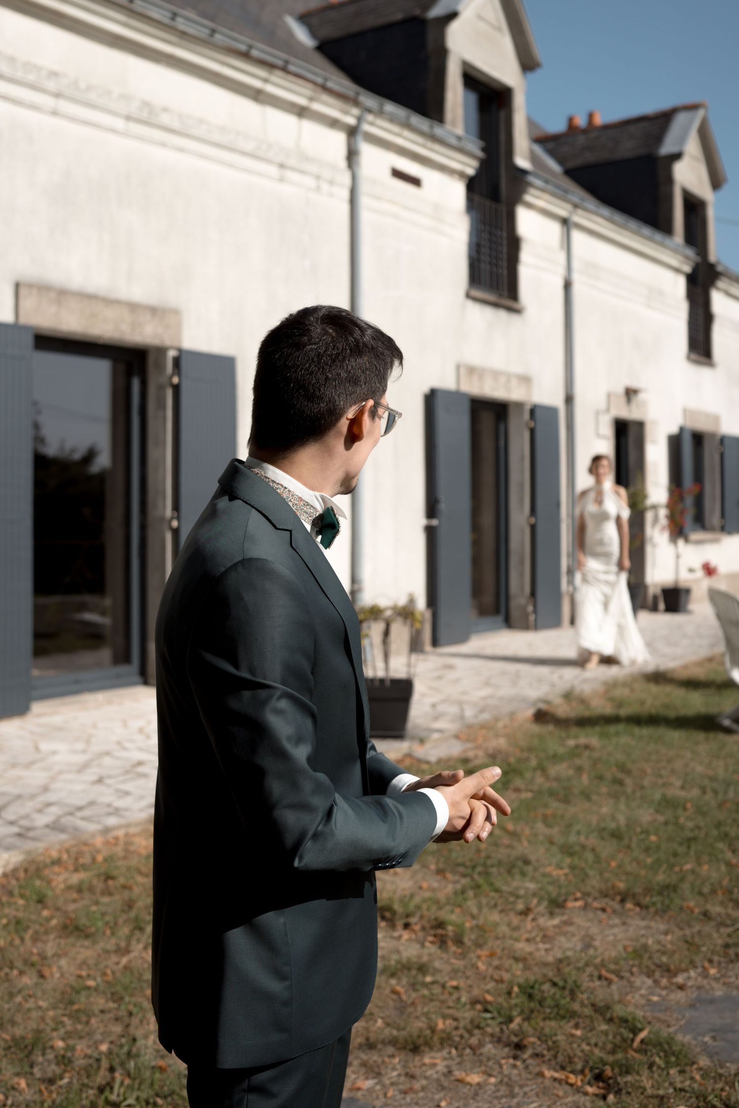 Le marié en costume sombre regarde la mariée s'approcher en robe blanche devant un élégant château blanc angers.