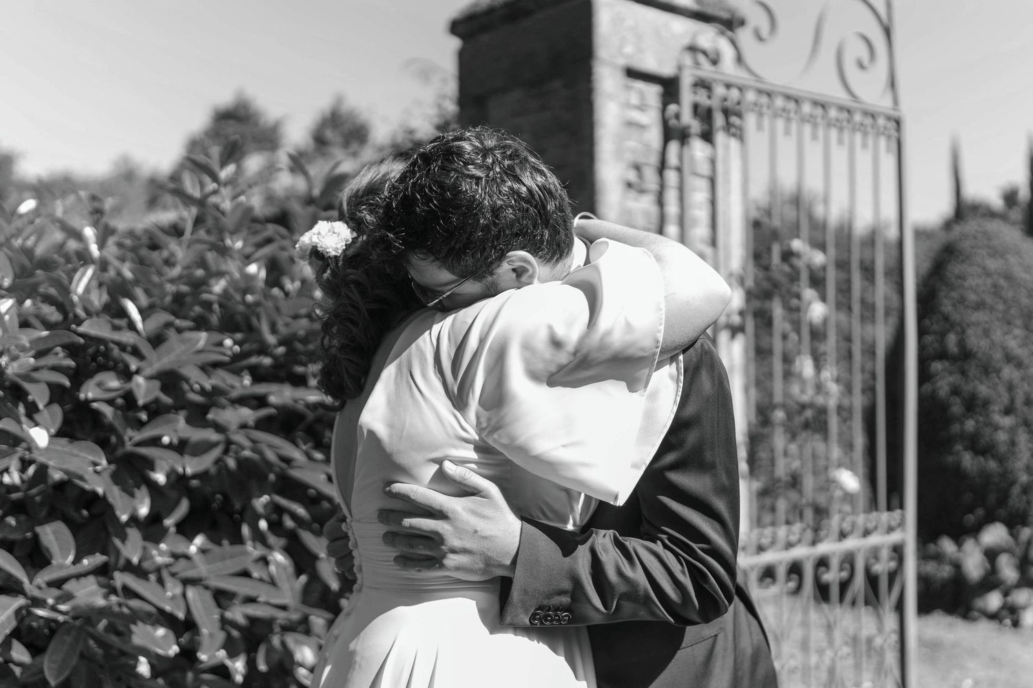 Étreinte des mariés en noir et blanc – Moment d’émotion capturé devant un portail en fer forgé et un jardin.