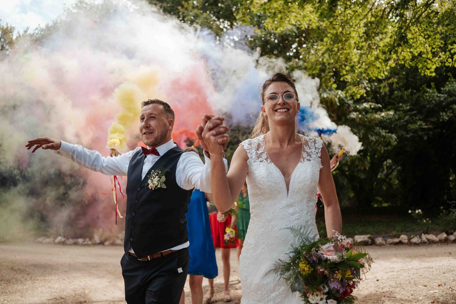 Les mariés, rayonnants de bonheur, traversent des fumigènes colorés lors d'une fête de mariage en plein air.