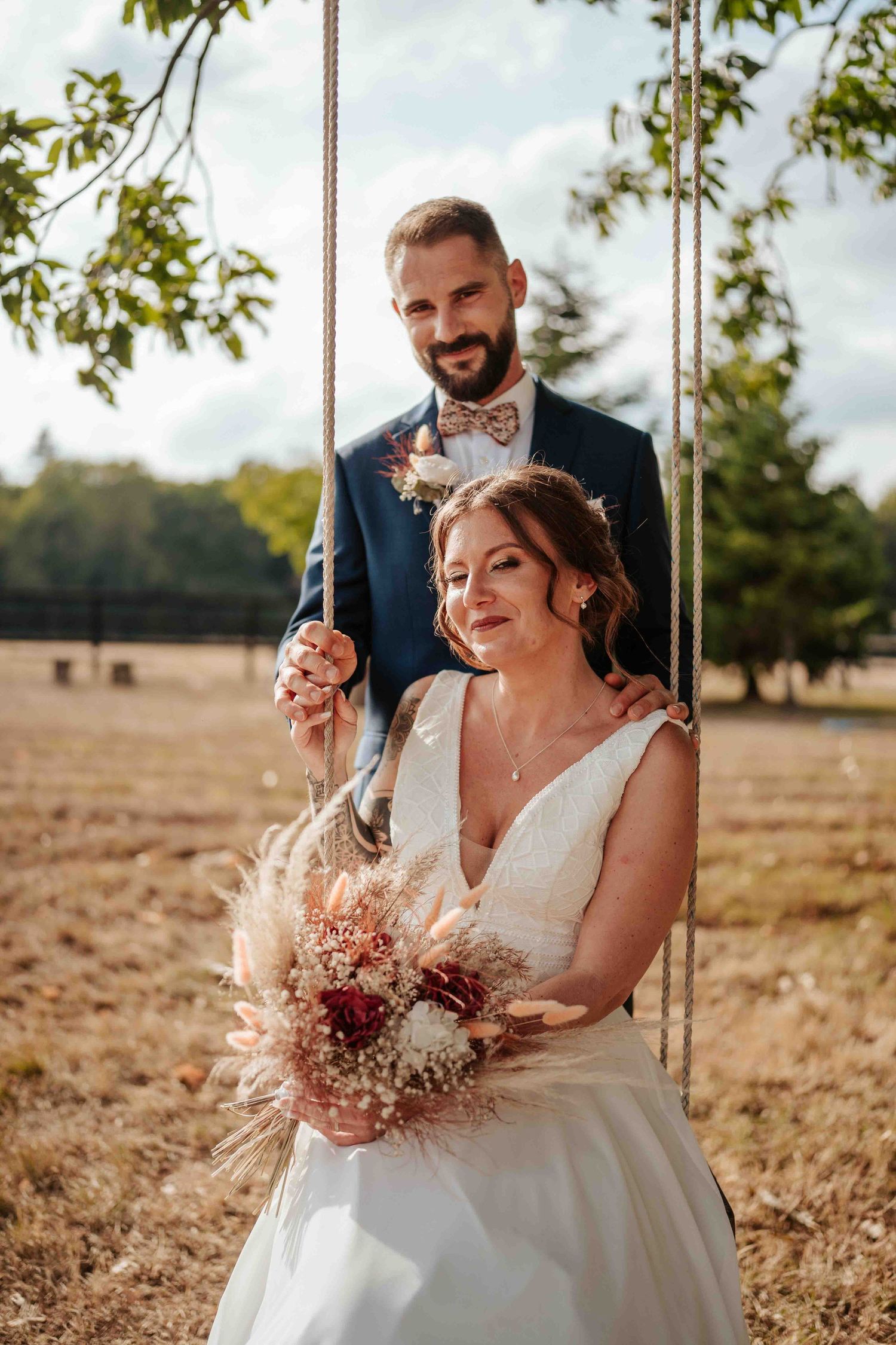 La mariée tient un bouquet de fleurs séchées, le marié se tenant derrière elle sur une balançoire en corde à l'extérieur.