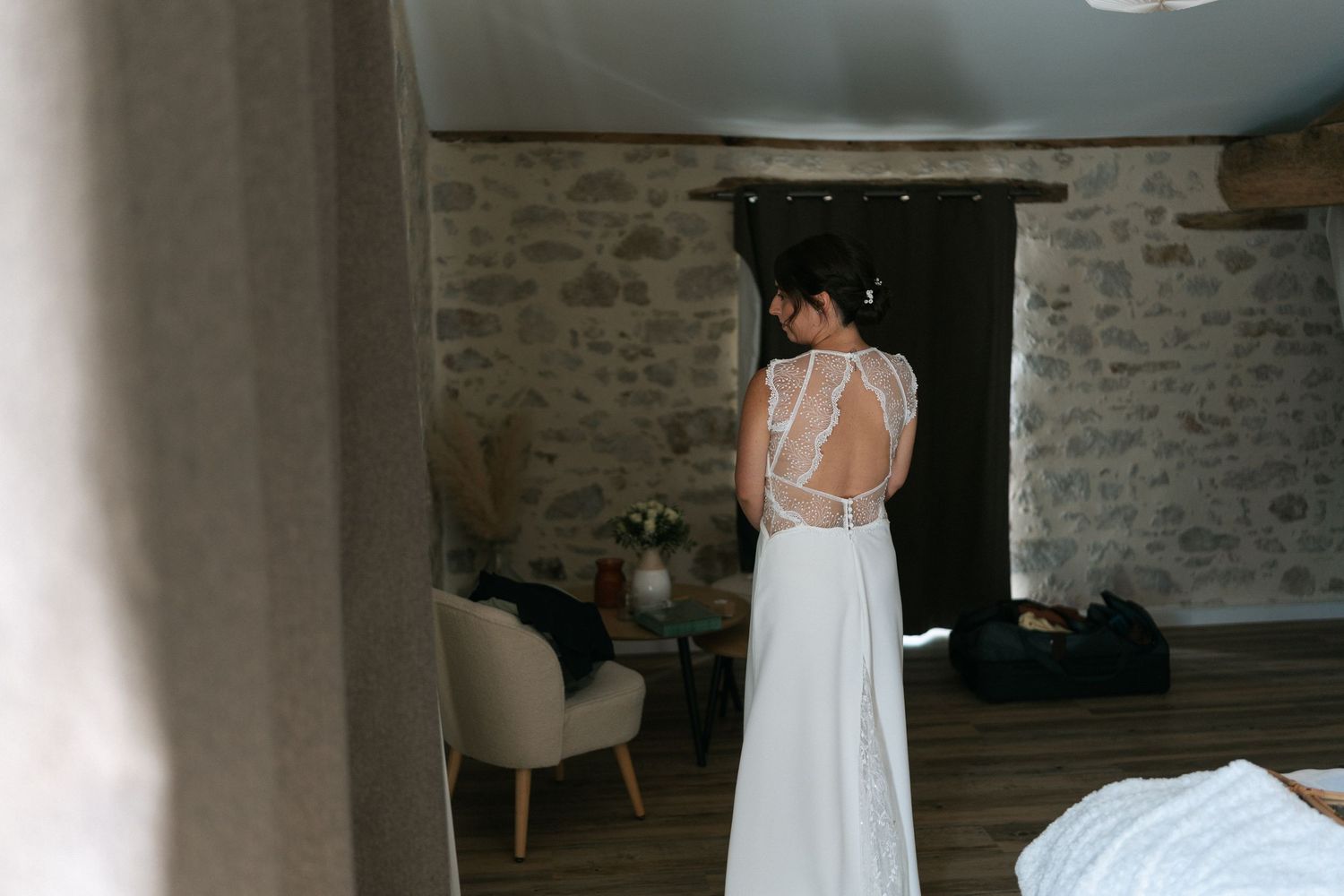 Bride in elegant backless wedding dress with lace detail stands in rustic stone-walled venue room.