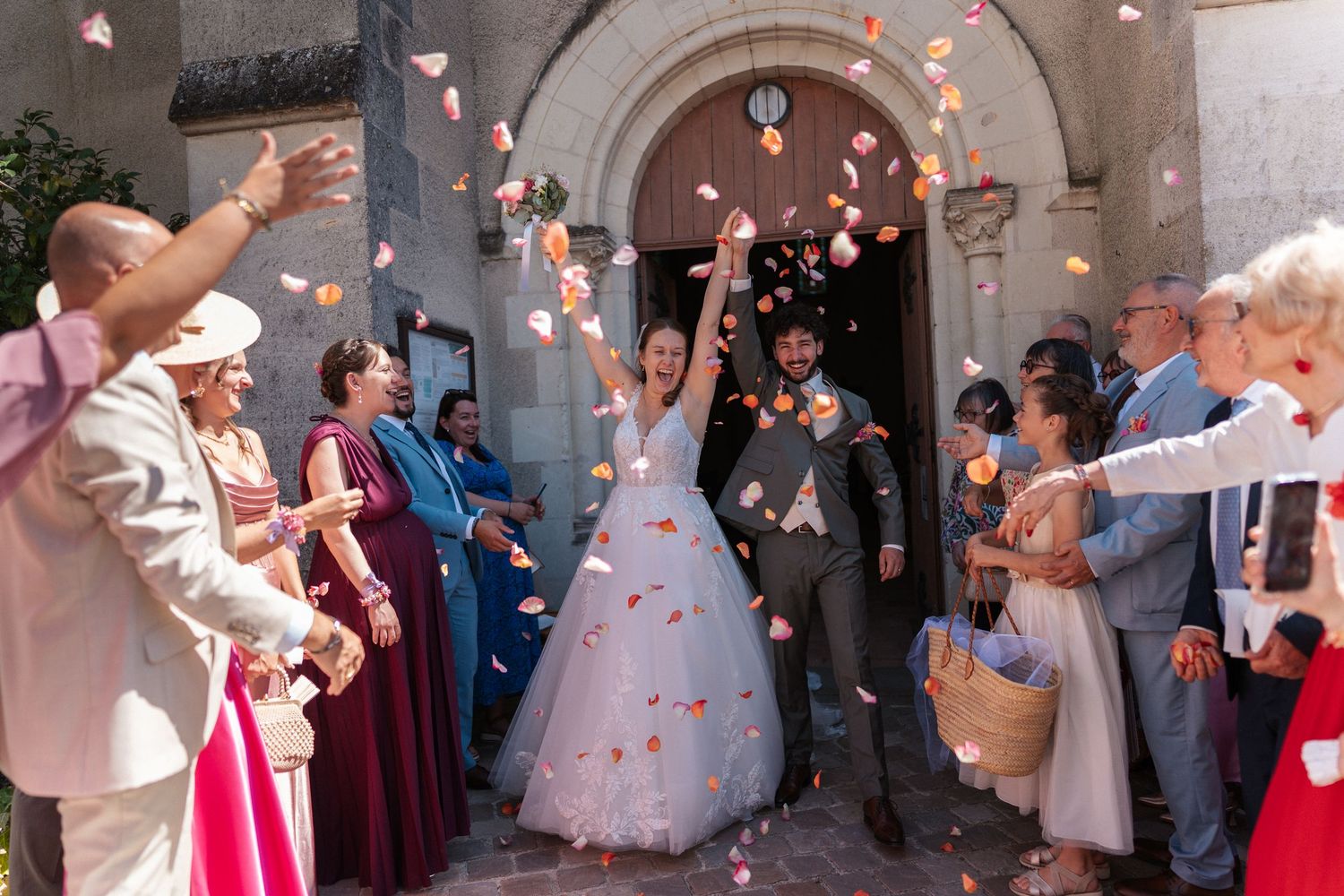 Bride and groom exit church amid flower petal confetti toss, surrounded by guests in formal attire.