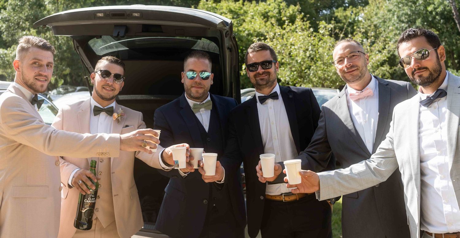 Un groupe de garçons d'honneur en costume trinquent avec des coupes à côté du coffre d'une voiture lors d'un mariage en plein air.