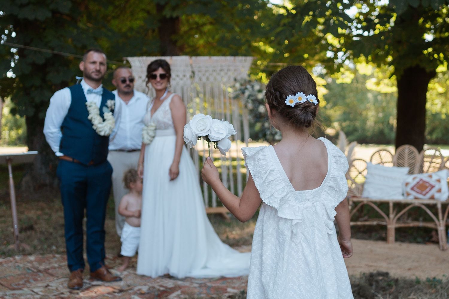 Mariés et enfants pendant une cérémonie laïque en extérieur – Petite fille tenant un bouquet face aux mariés devant une arche décorée.
