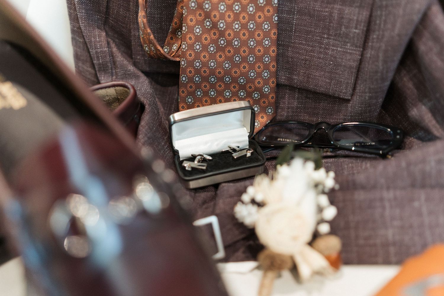 Un costume en tweed marron avec cravate à motifs, boutons de manchette dans leur boîte, lunettes de soleil et boutonnière blanche.