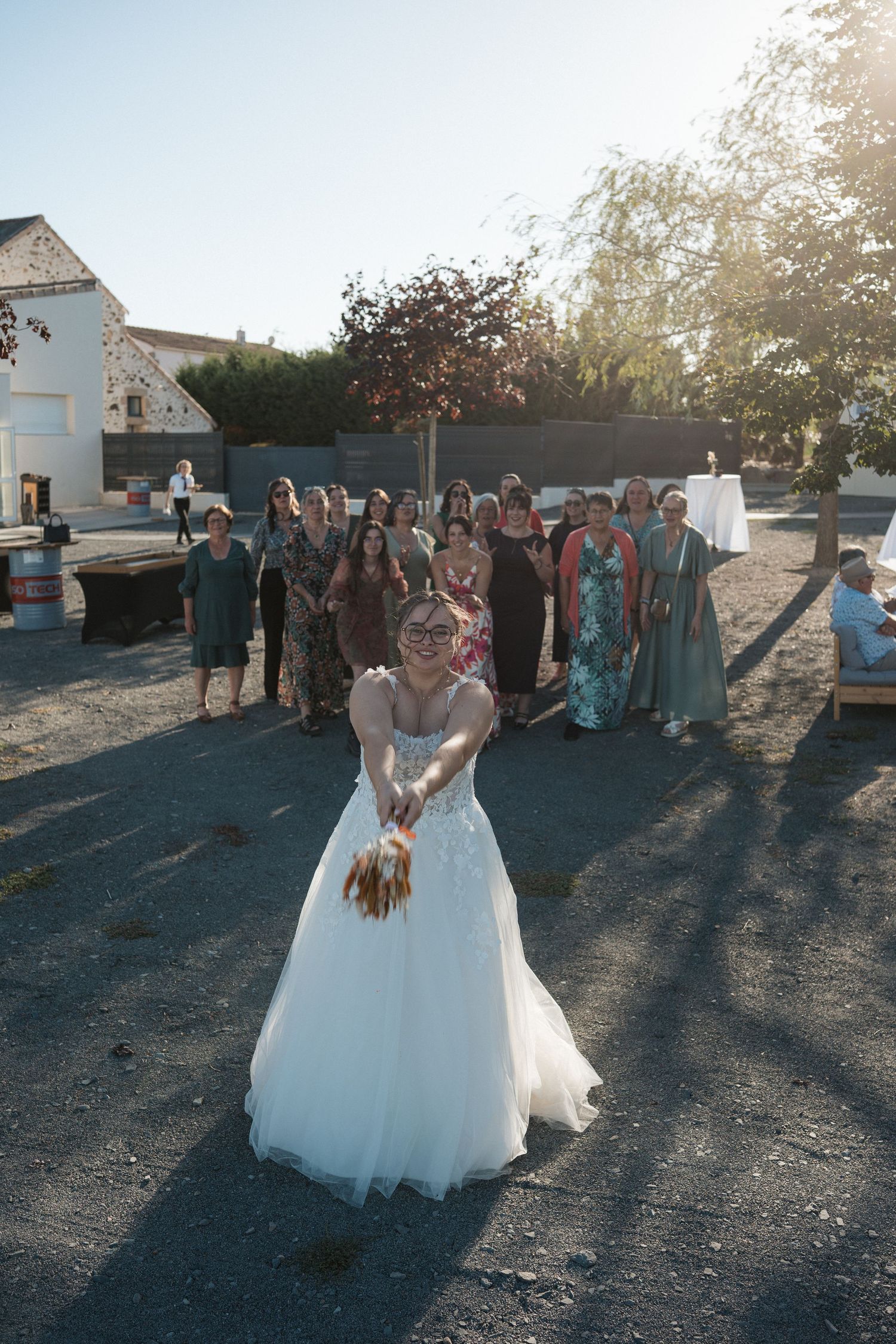 Bride in white gown tosses bouquet to guests gathered outdoors at sunny wedding reception.