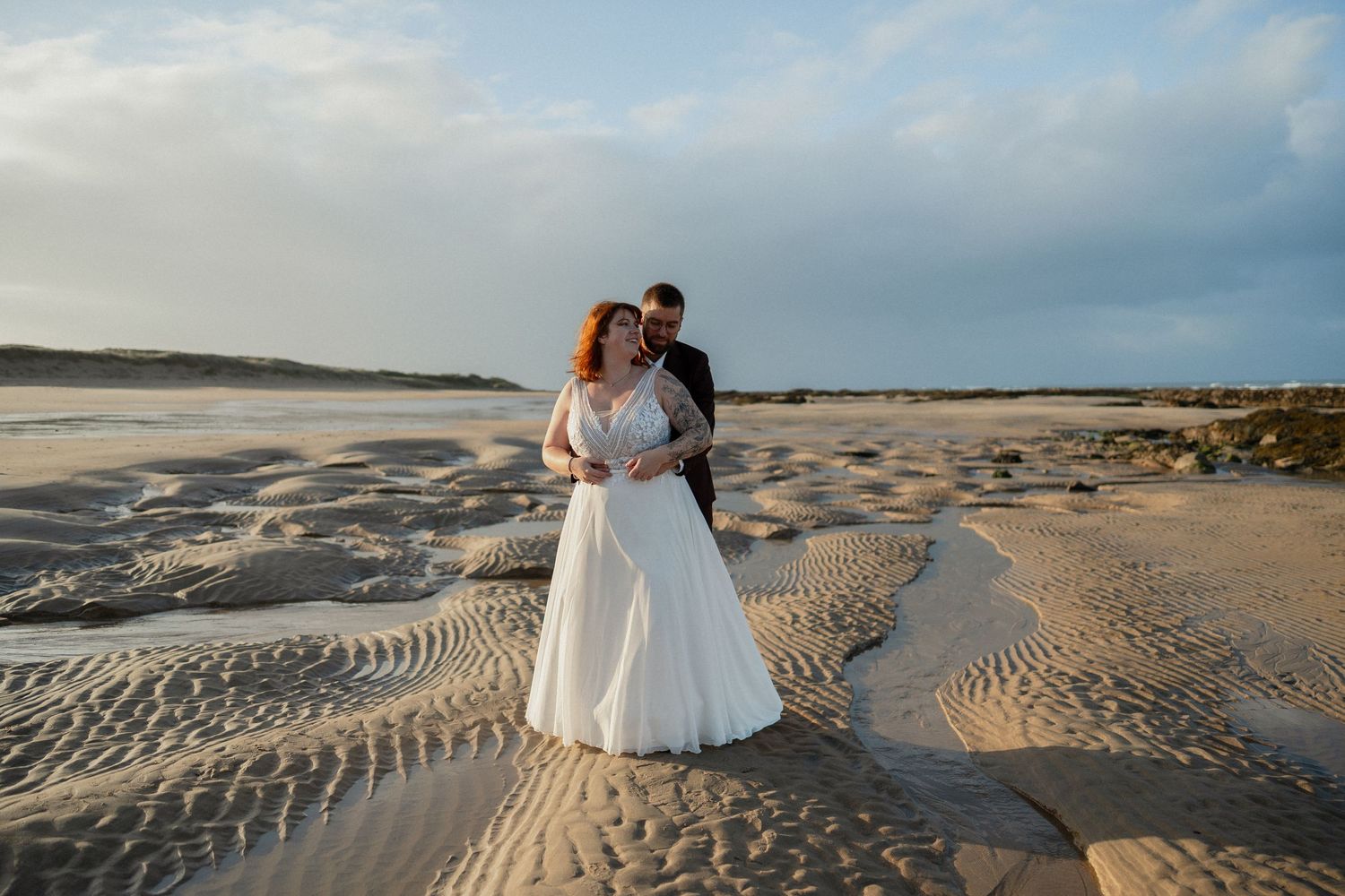 Les mariés s'enlacent sur une plage de sable fin à l'heure dorée, avec les vagues de l'océan et un ciel spectaculaire en arrière-plan.
