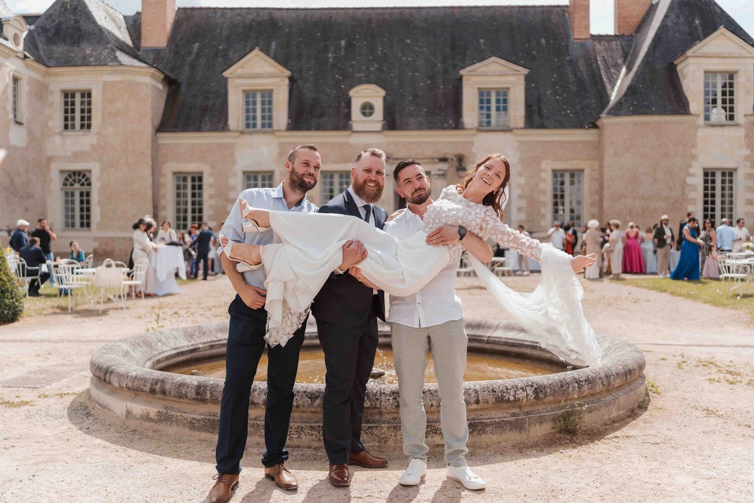 Trois garçons d'honneur soulèvent joyeusement les mariés devant un château français avec fontaine, lieu de réception de mariage.