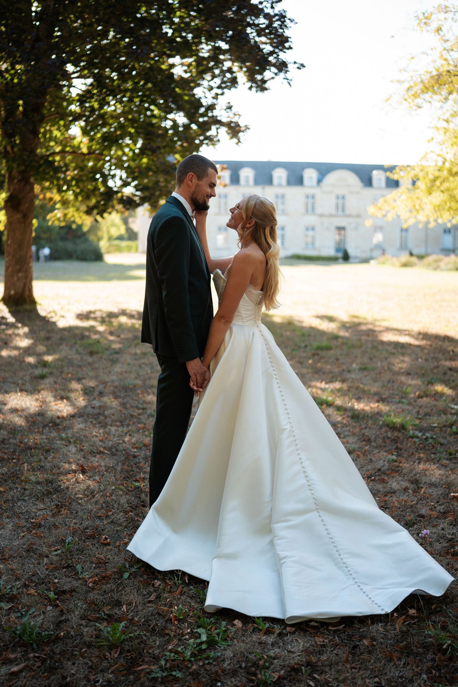 Bride in white ball gown and groom in dark suit hold hands outdoors near a French chateau at golden hour.