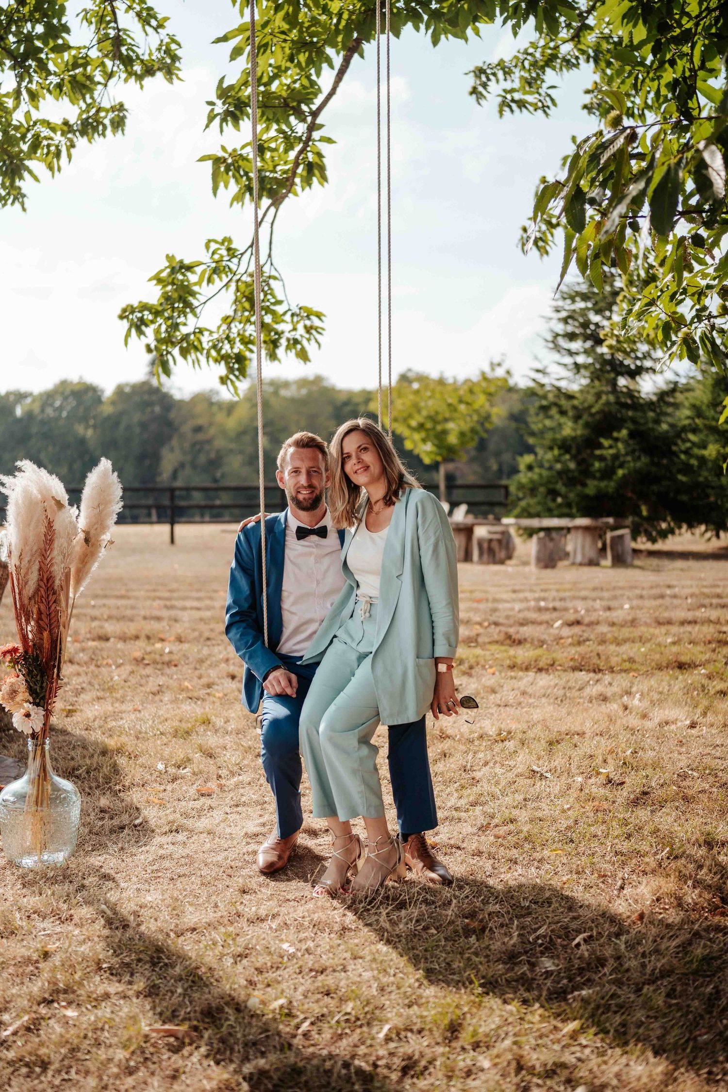 Un couple en costumes élégants pose sur une balançoire en corde dans un champ ensoleillé à l'occasion de leur mariage.
