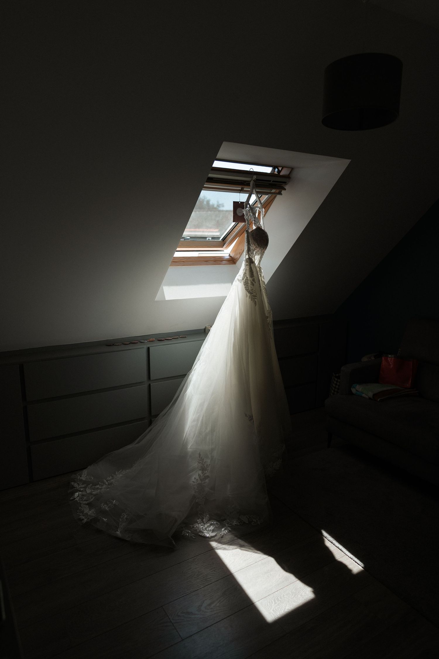 Elegant wedding dress hanging from attic skylight window, casting dramatic shadows in dimly lit room.