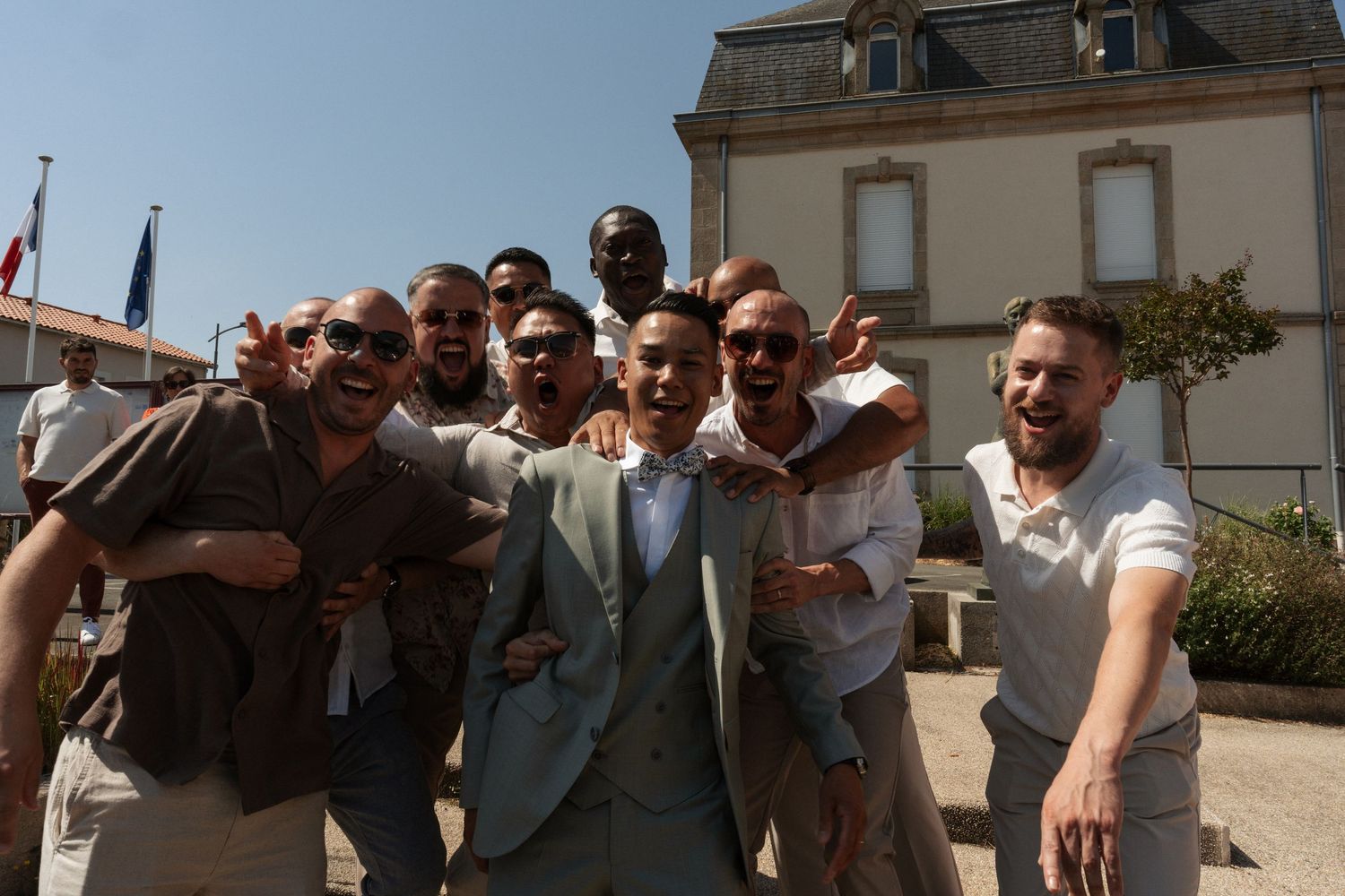 Group of men smiling and posing together outdoors in front of a building on a sunny day.