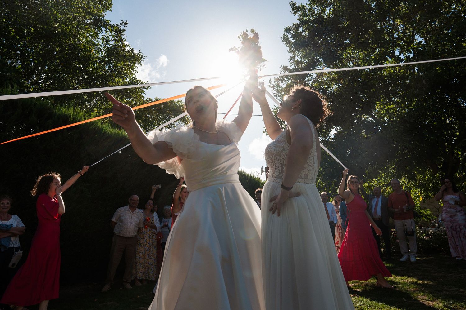 Two brides in white gowns celebrate outdoors at a same-sex wedding ceremony under sunlit trees with guests watching.