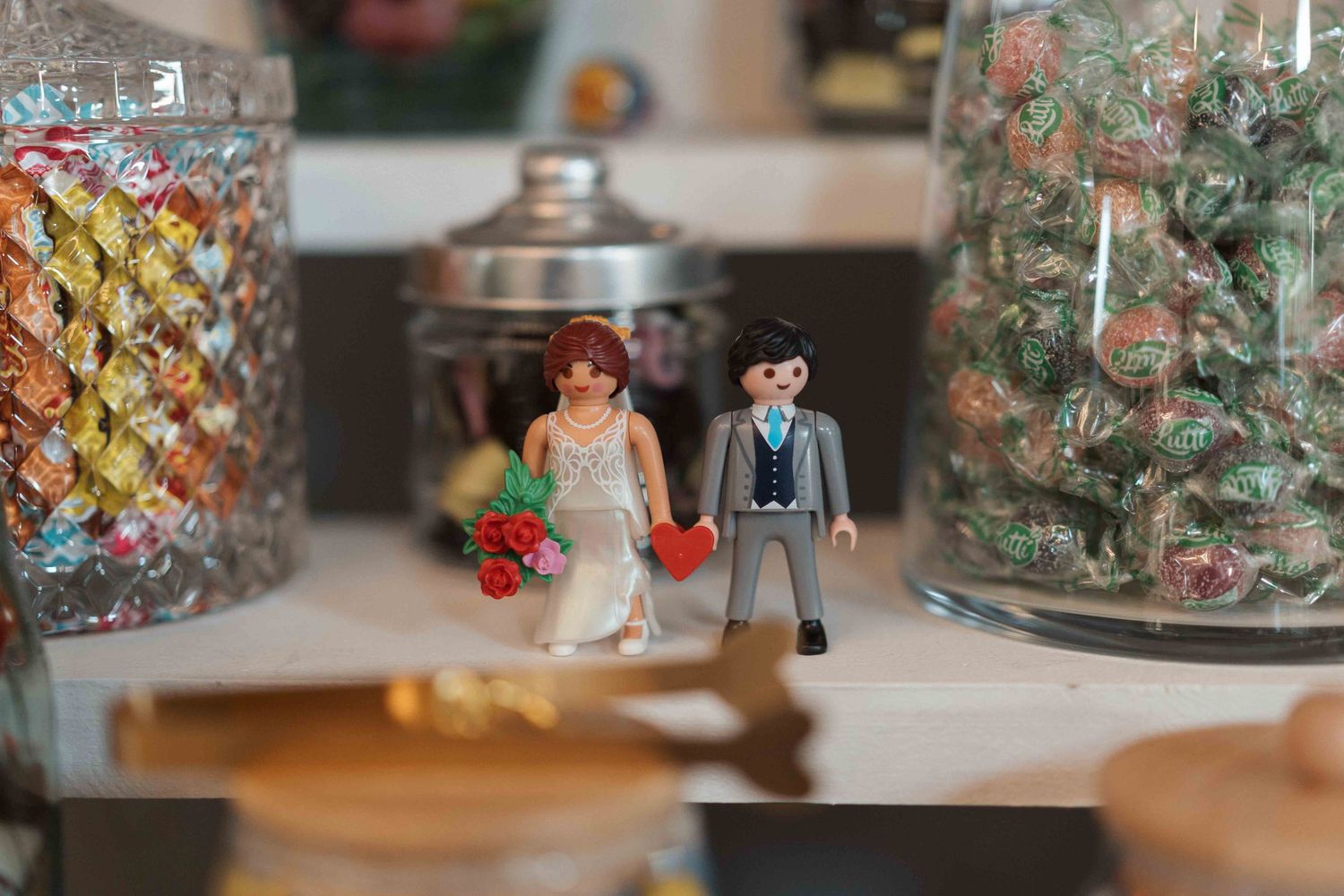 Figurines Playmobil de couple de mariés présentées entre des bocaux en verre remplis de bonbons colorés sur une surface blanche.