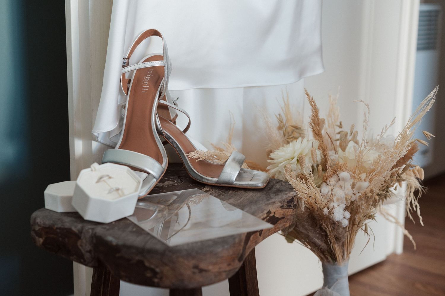 Escarpins argentés à brides pour une mariée, posés sur un tabouret rustique avec une boîte à alliances et un bouquet de fleurs séchées.