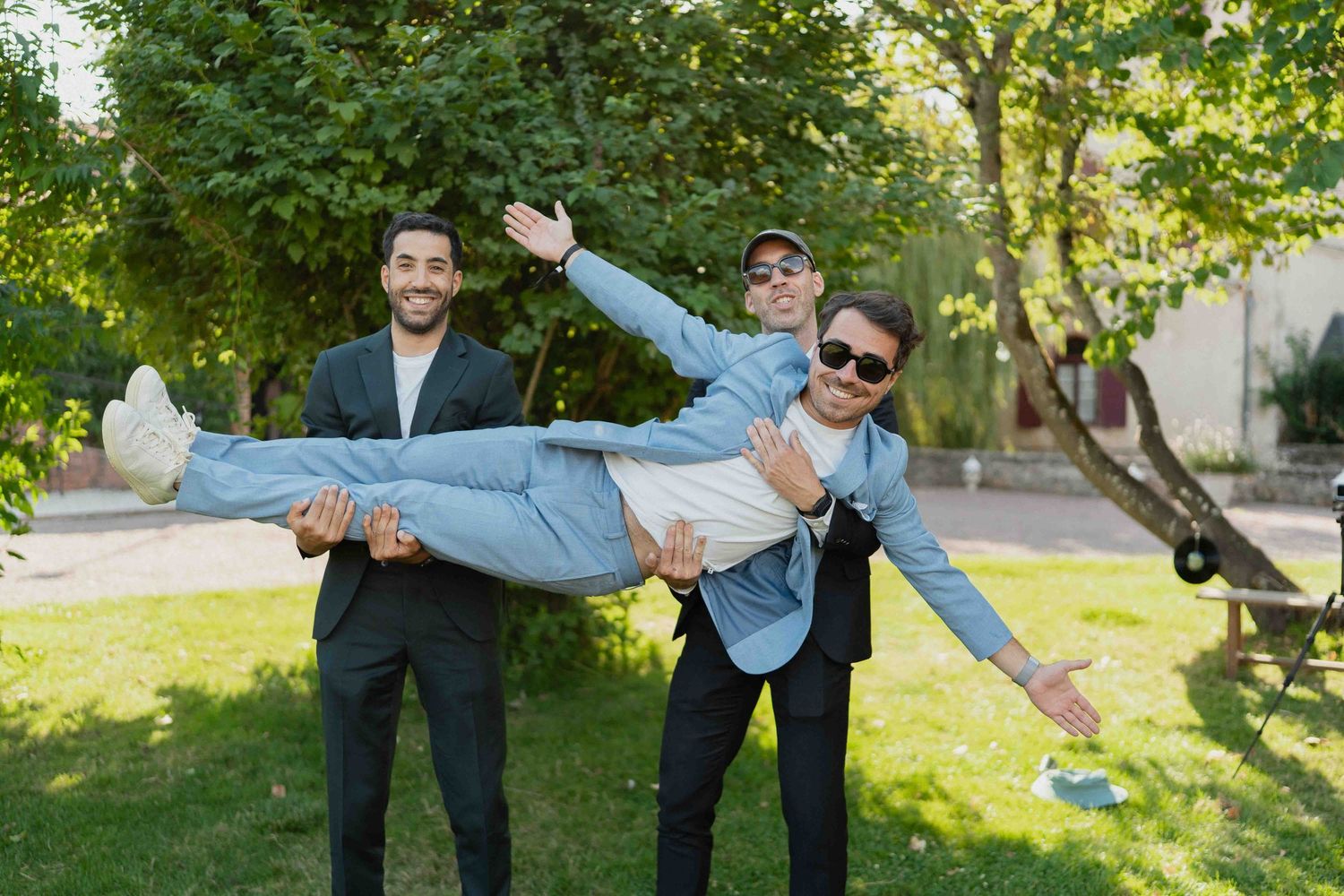 Trois jeunes hommes soulèvent un ami à l'horizontale en plaisantant, dans un jardin verdoyant.