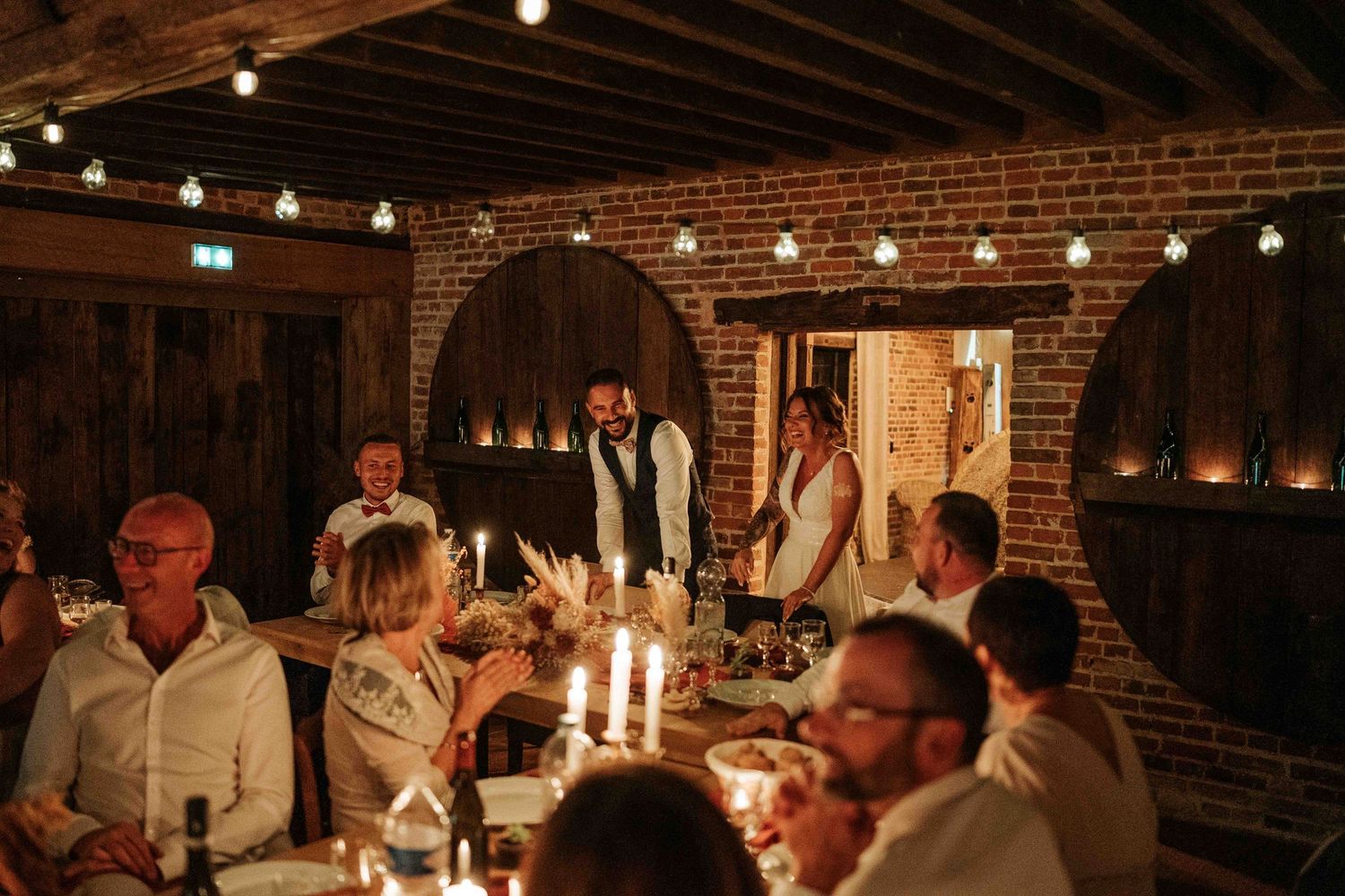 Un couple prononçait un discours lors d'une réception de mariage champêtre éclairée aux chandelles dans une grange en briques.