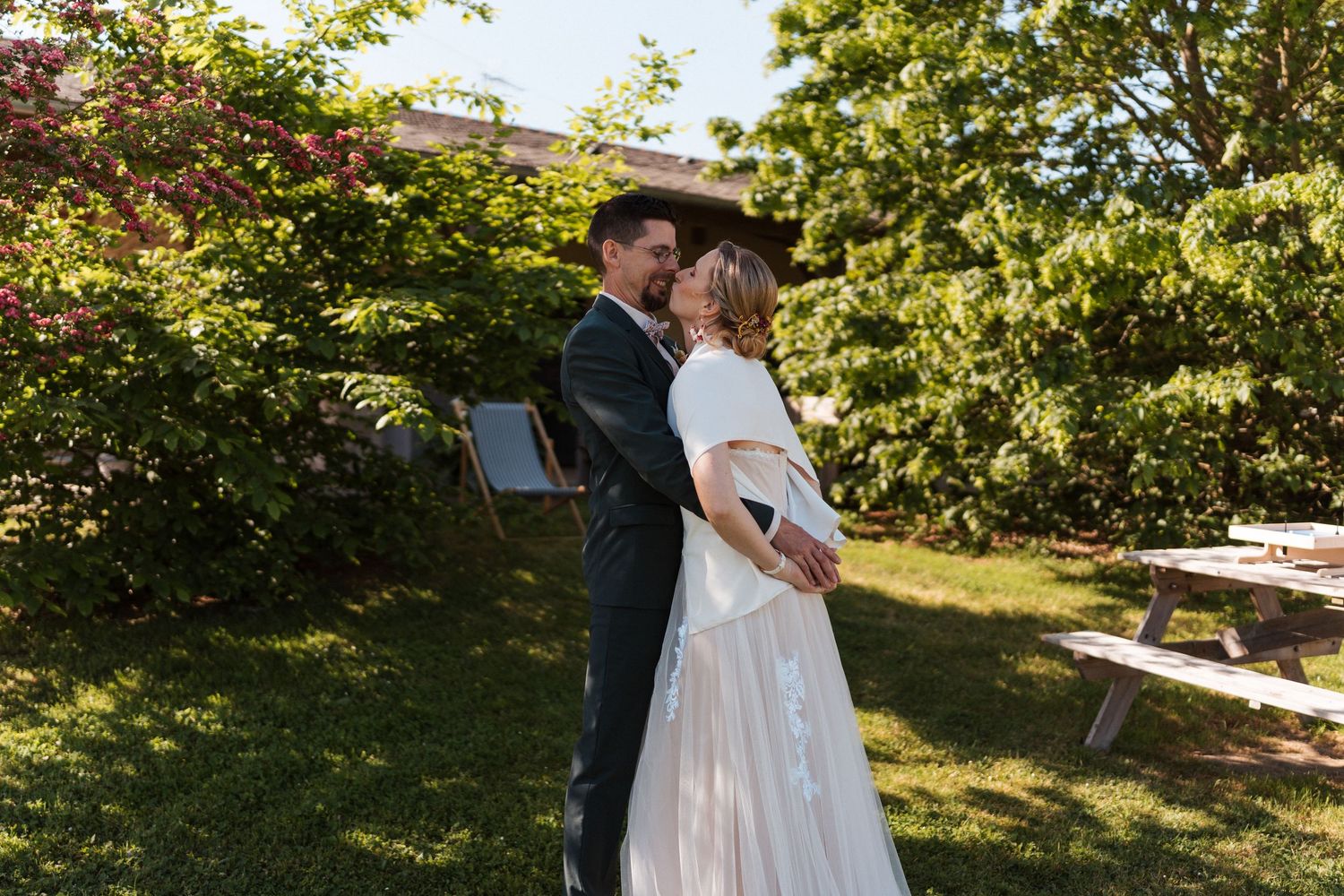 A bride and groom share a romantic moment outdoors on a sunny day, surrounded by lush green trees and garden scenery.