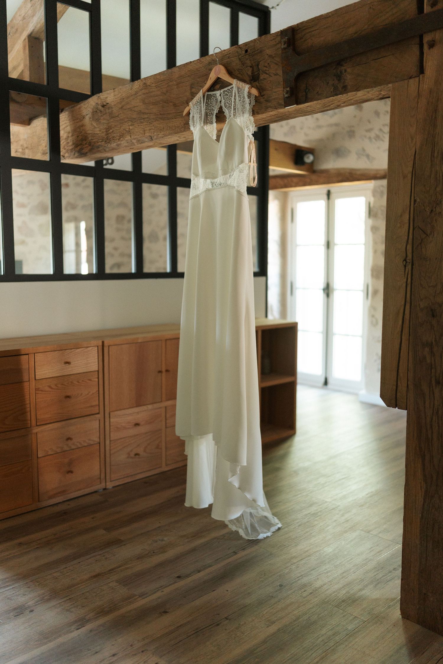 Elegant white wedding dress hanging on a wooden beam in a rustic room with hardwood floors and wooden furniture.