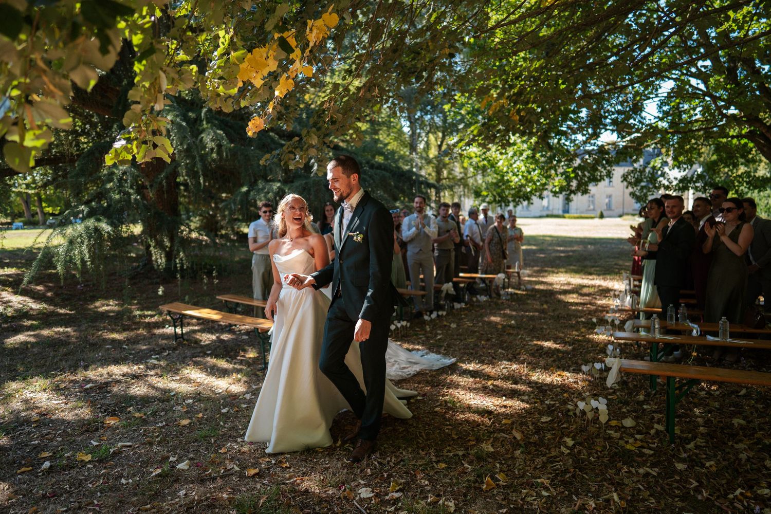 Bride and groom walk down outdoor aisle under trees at a sunny garden wedding ceremony.