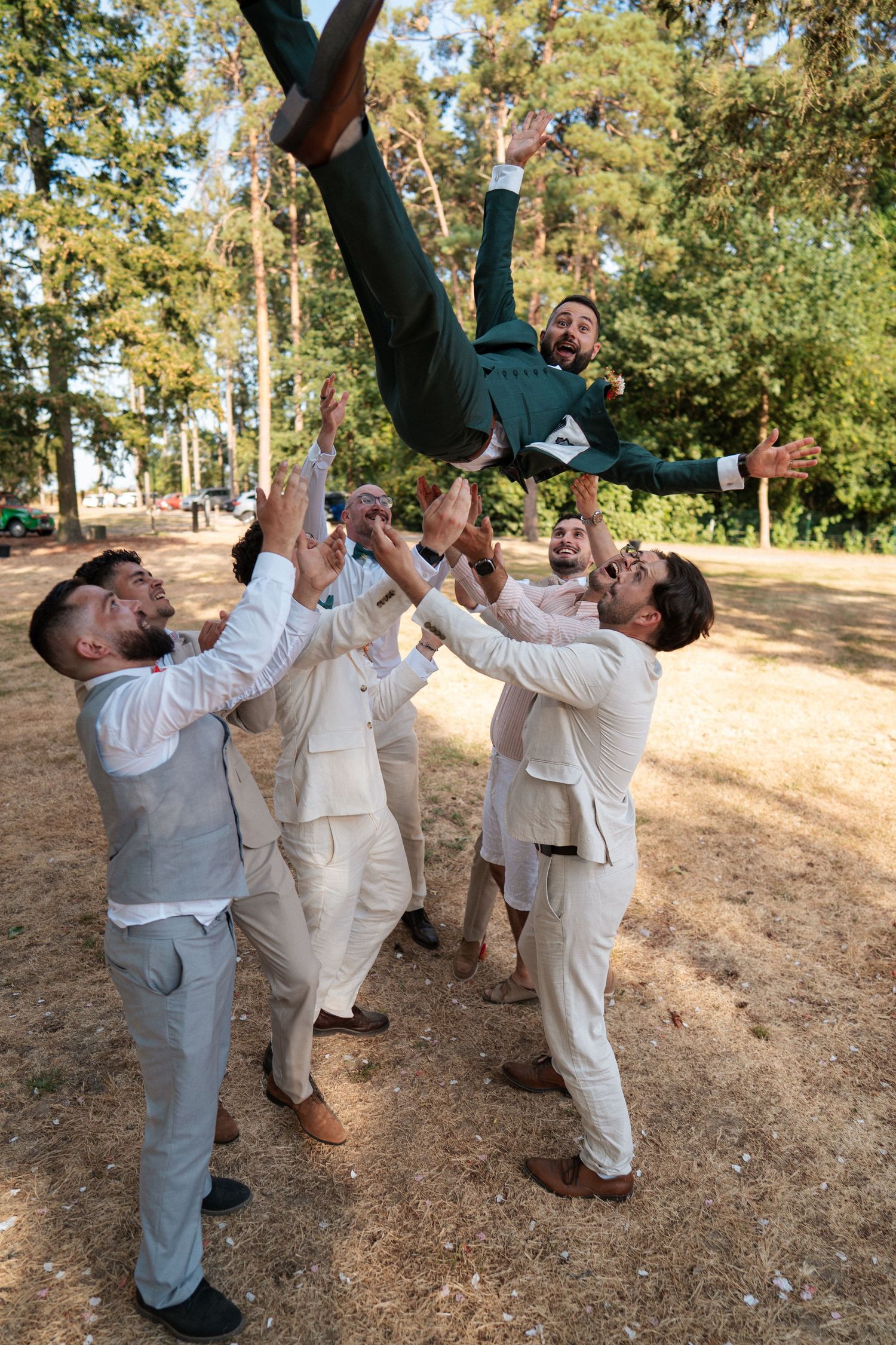 Groomsmen tossing groom in the air outdoors during a wedding celebration in a sunny park.