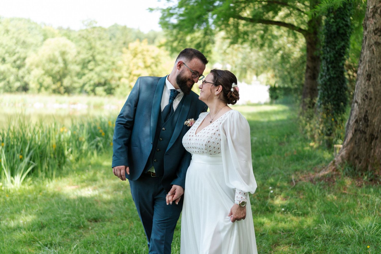 A bride and groom share a romantic moment outdoors near a pond, surrounded by lush greenery on their wedding day.