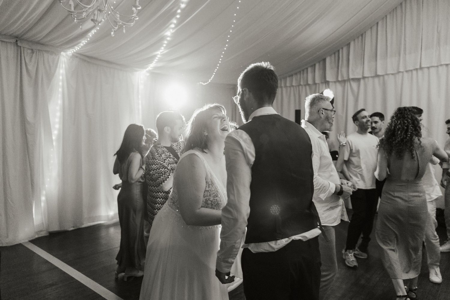Soirée de mariage en noir et blanc sous chapiteau – Mariés et invités dansant sous des guirlandes lumineuses.
