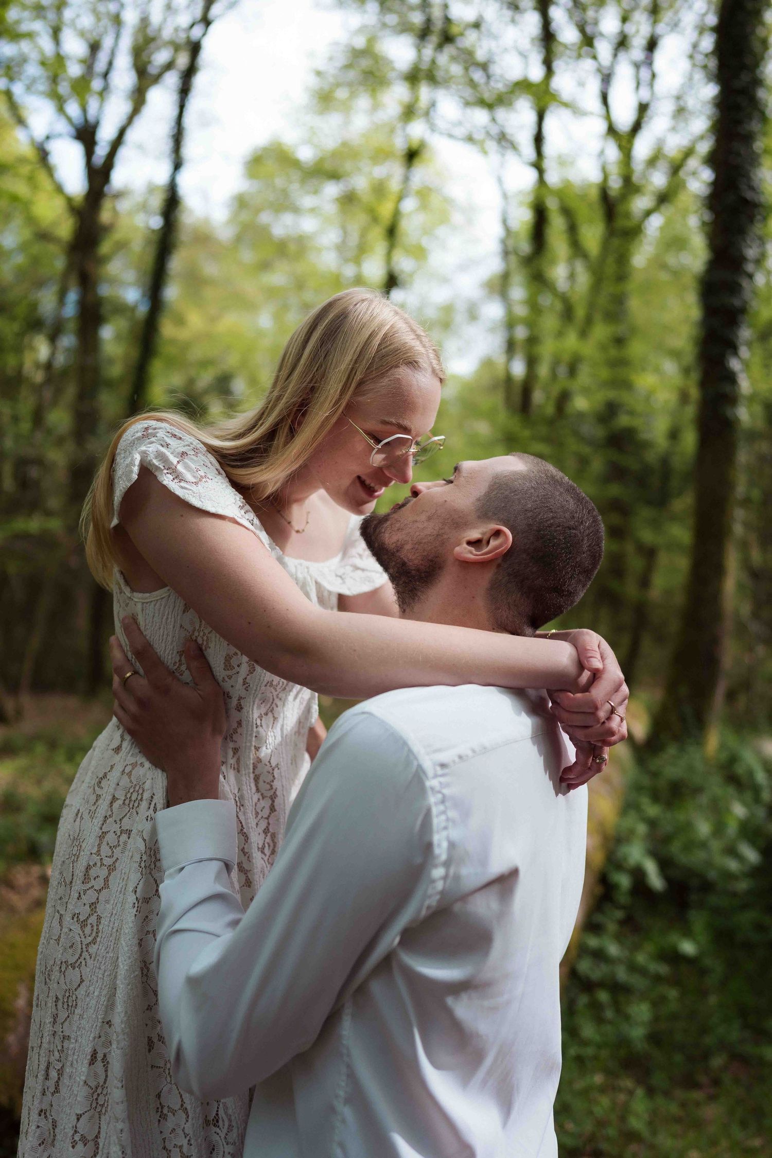 Un couple s'enlace joyeusement dans une forêt ensoleillée, la femme en robe de dentelle soulevée par son partenaire.