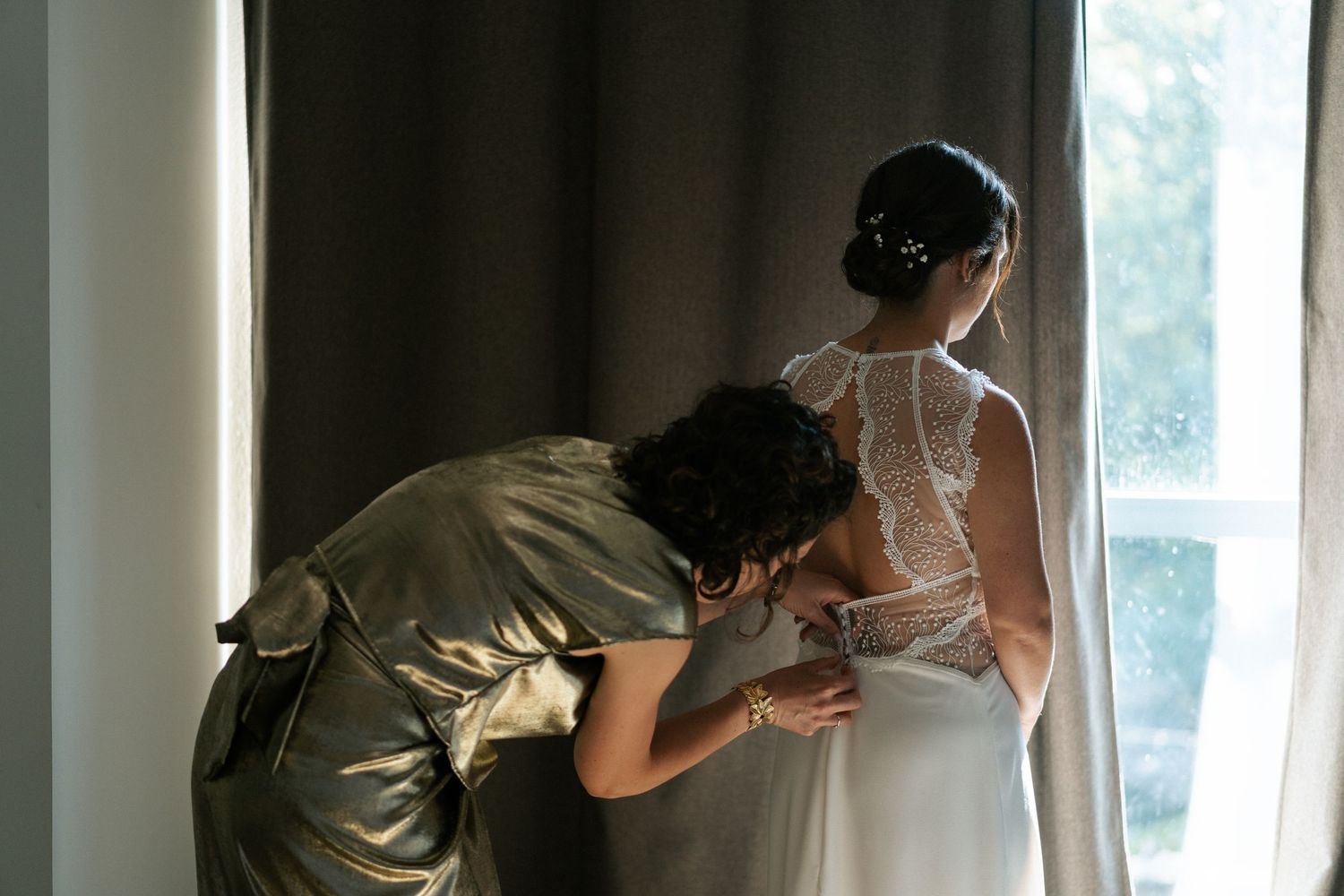 Bridesmaid in gold dress fastening the back of a bride's lace wedding gown near a window.