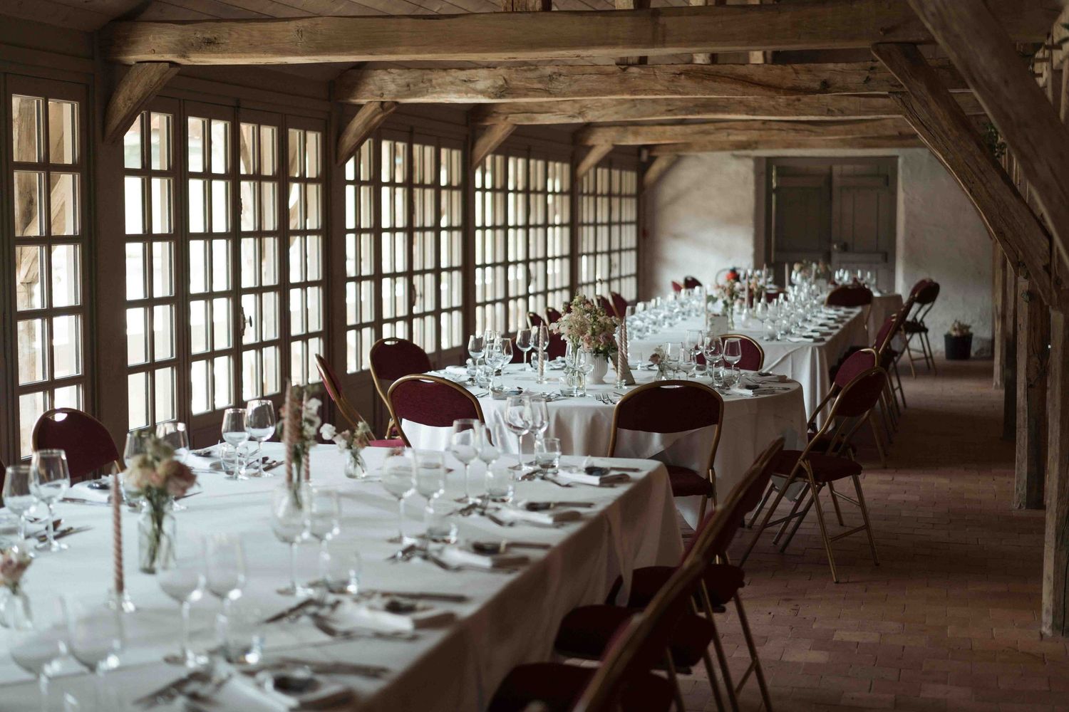 De longues tables de banquet élégamment dressées, ornées de verrerie et de centres de table floraux, dans une grange rustique.