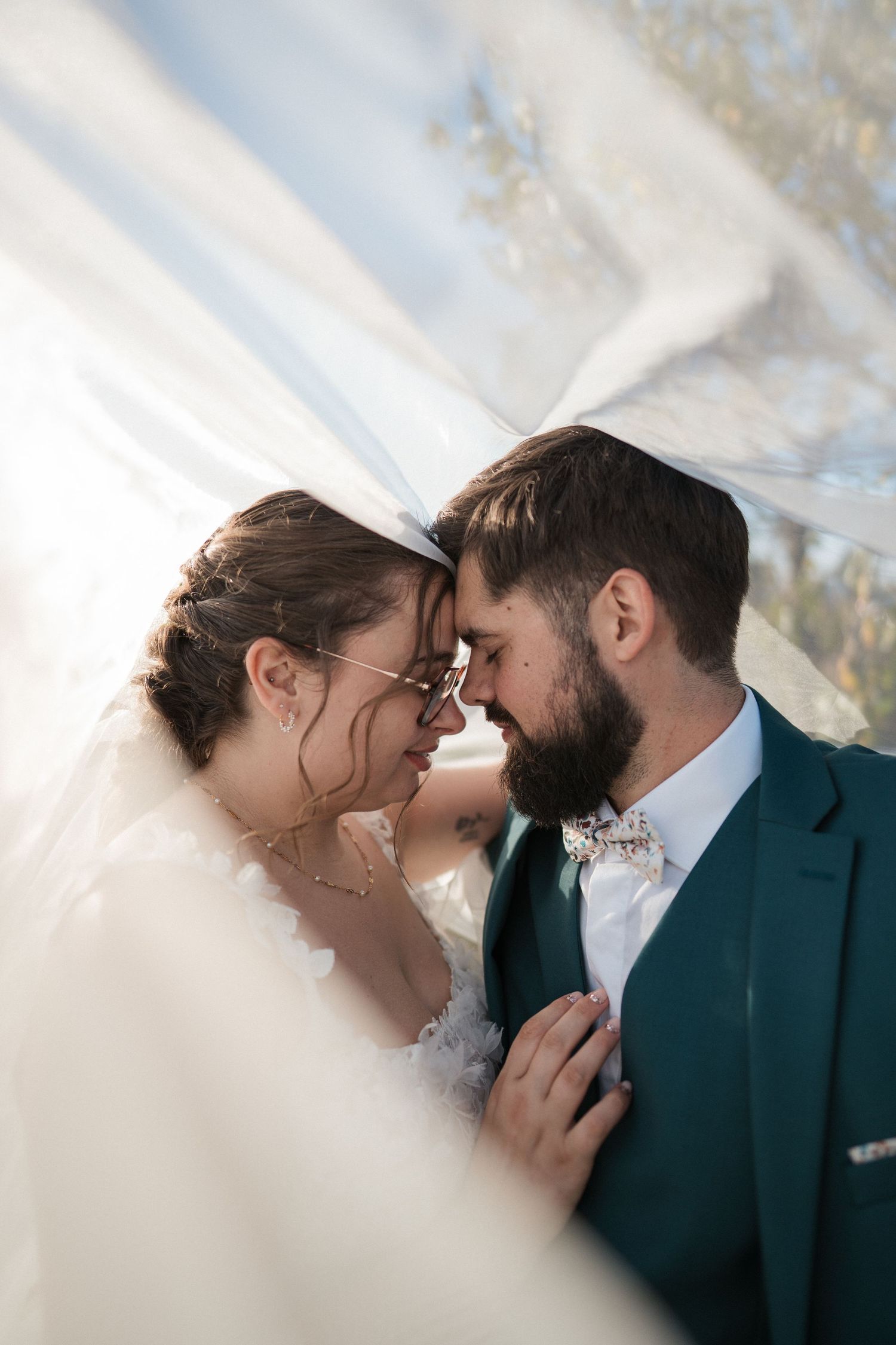 Bride and groom touching foreheads under flowing veil, groom in teal suit with floral bow tie.