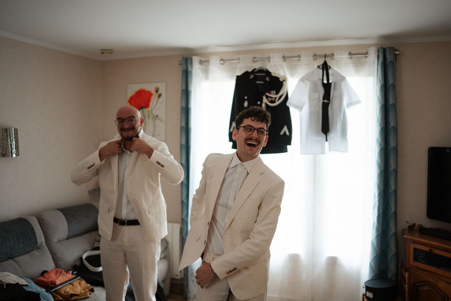 Two men in white suits getting ready indoors, adjusting ties with formal attire hanging in the background.