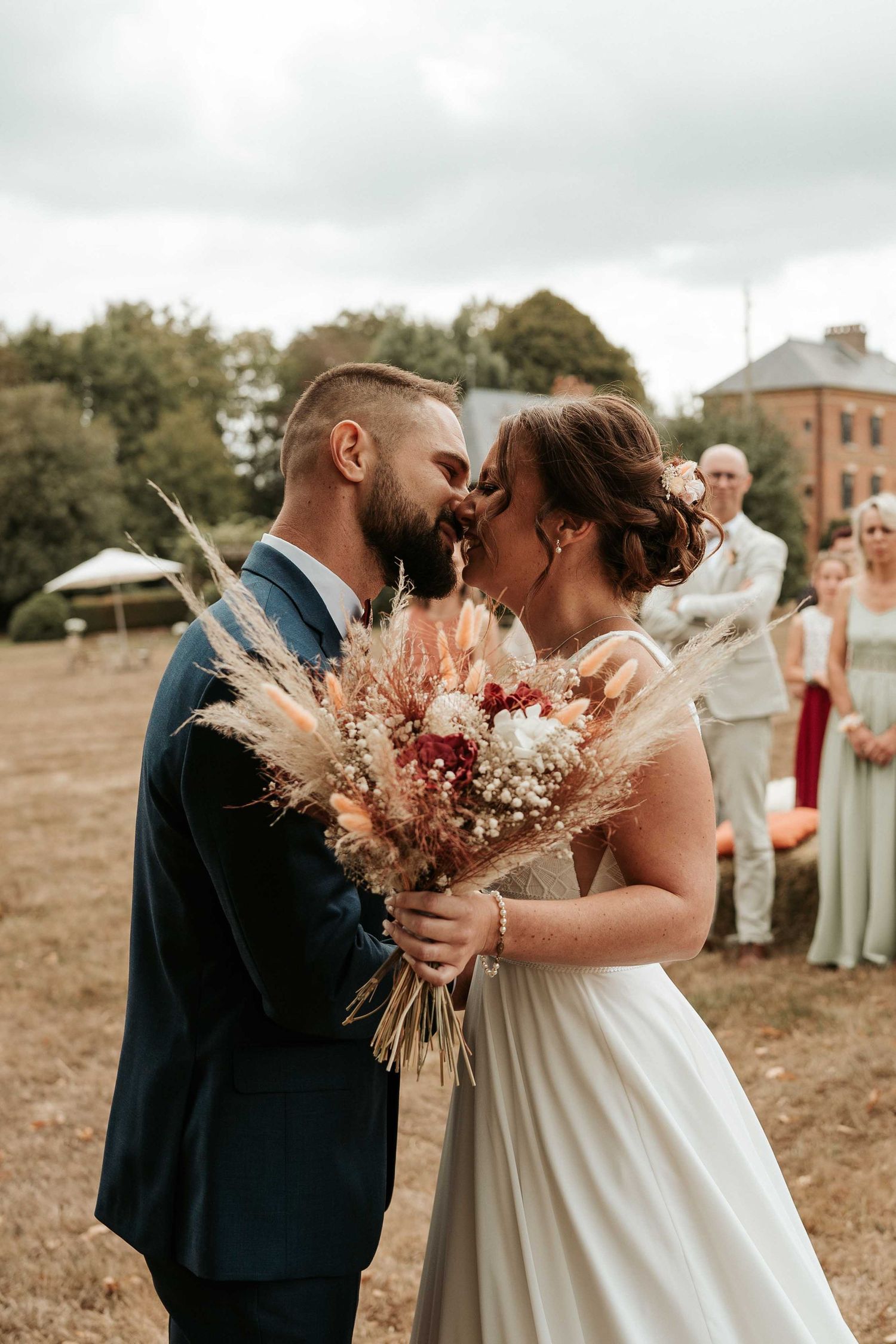 Les mariés échangent un baiser en tenant un bouquet de fleurs séchées de style bohème lors d'une cérémonie de mariage champêtre en extérieur