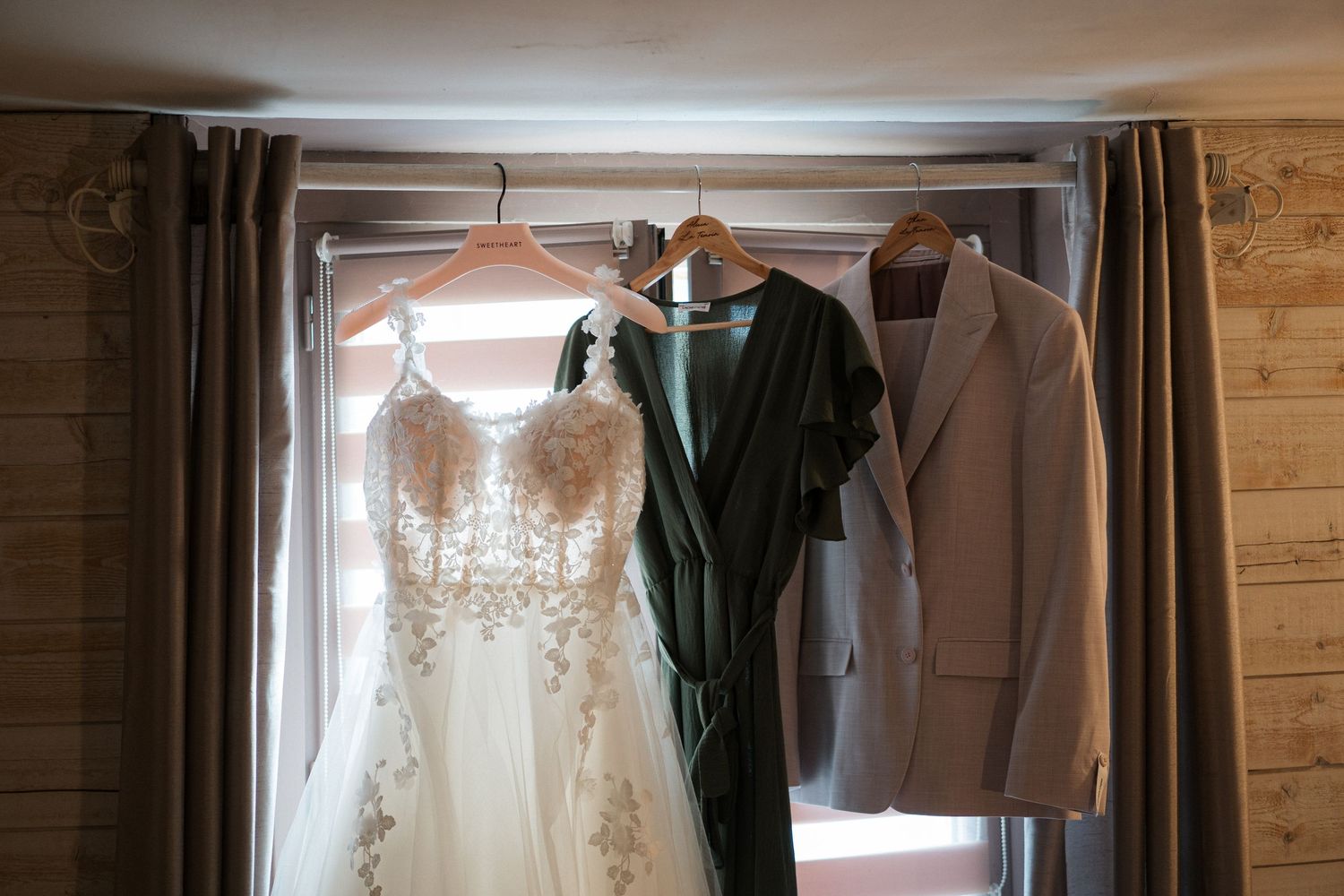Wedding dress, dark robe, and beige suit jacket hanging on wooden hangers in front of a window.