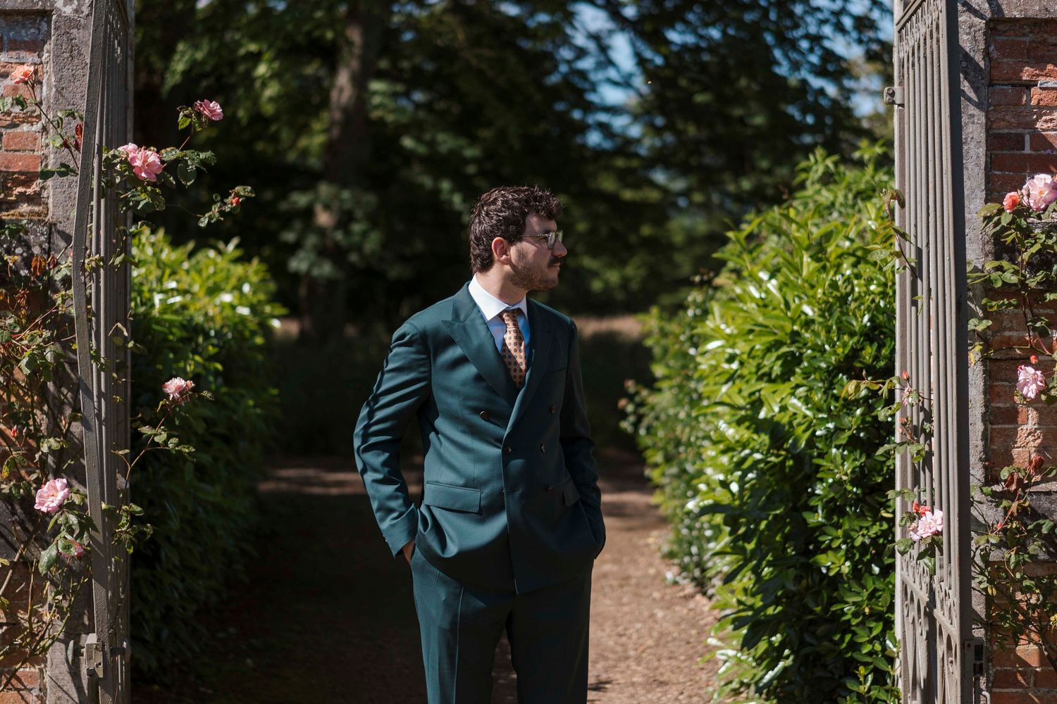 Un marié élégant, vêtu d'un costume turquoise et d'une cravate dorée, se tient sous une arche de jardin ensoleillée, entouré de roses.