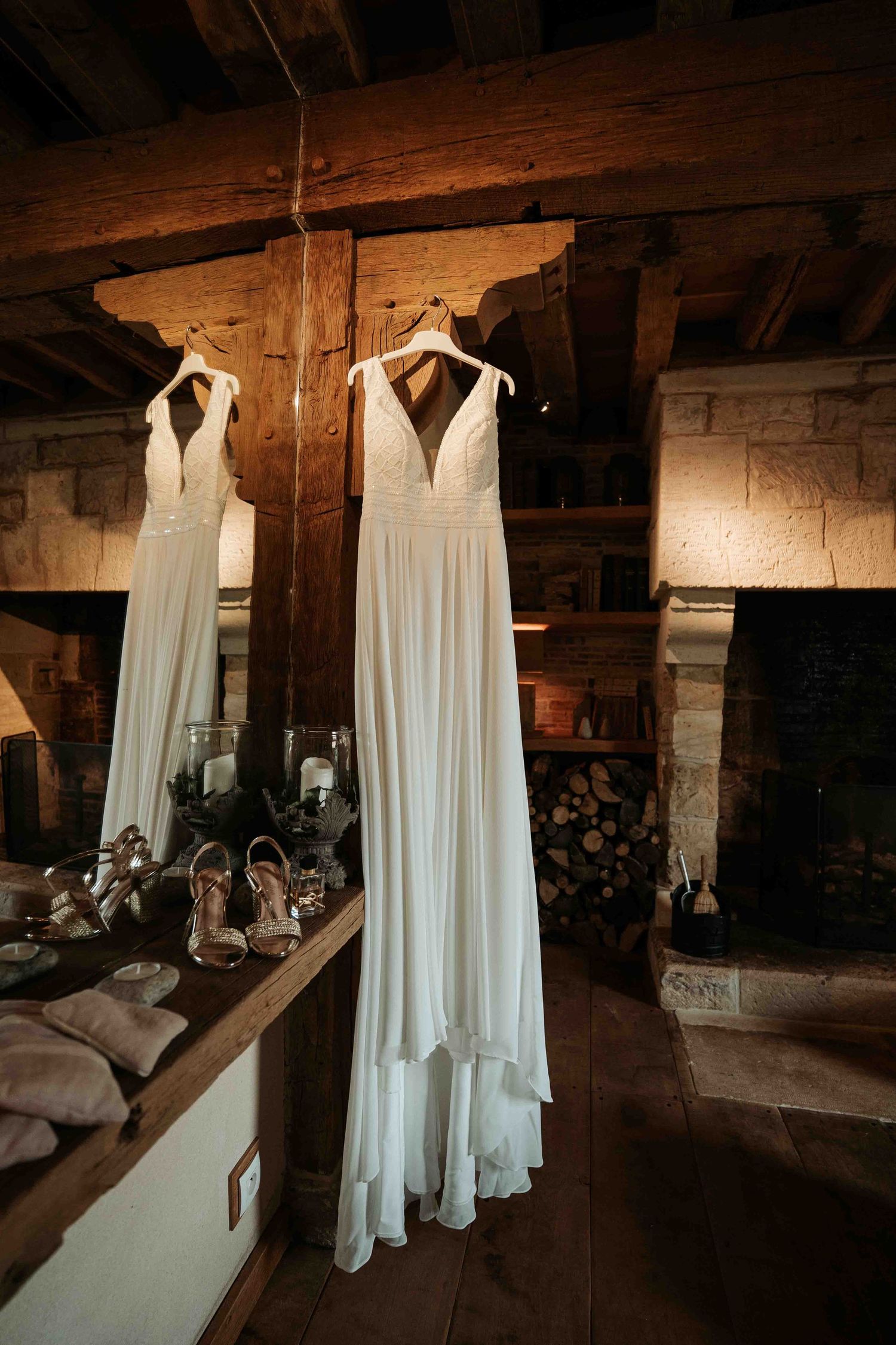 Robe de mariée blanche suspendue à des poutres en bois au-dessus d'un miroir dans une salle de réception rustique avec cheminée en pierre.