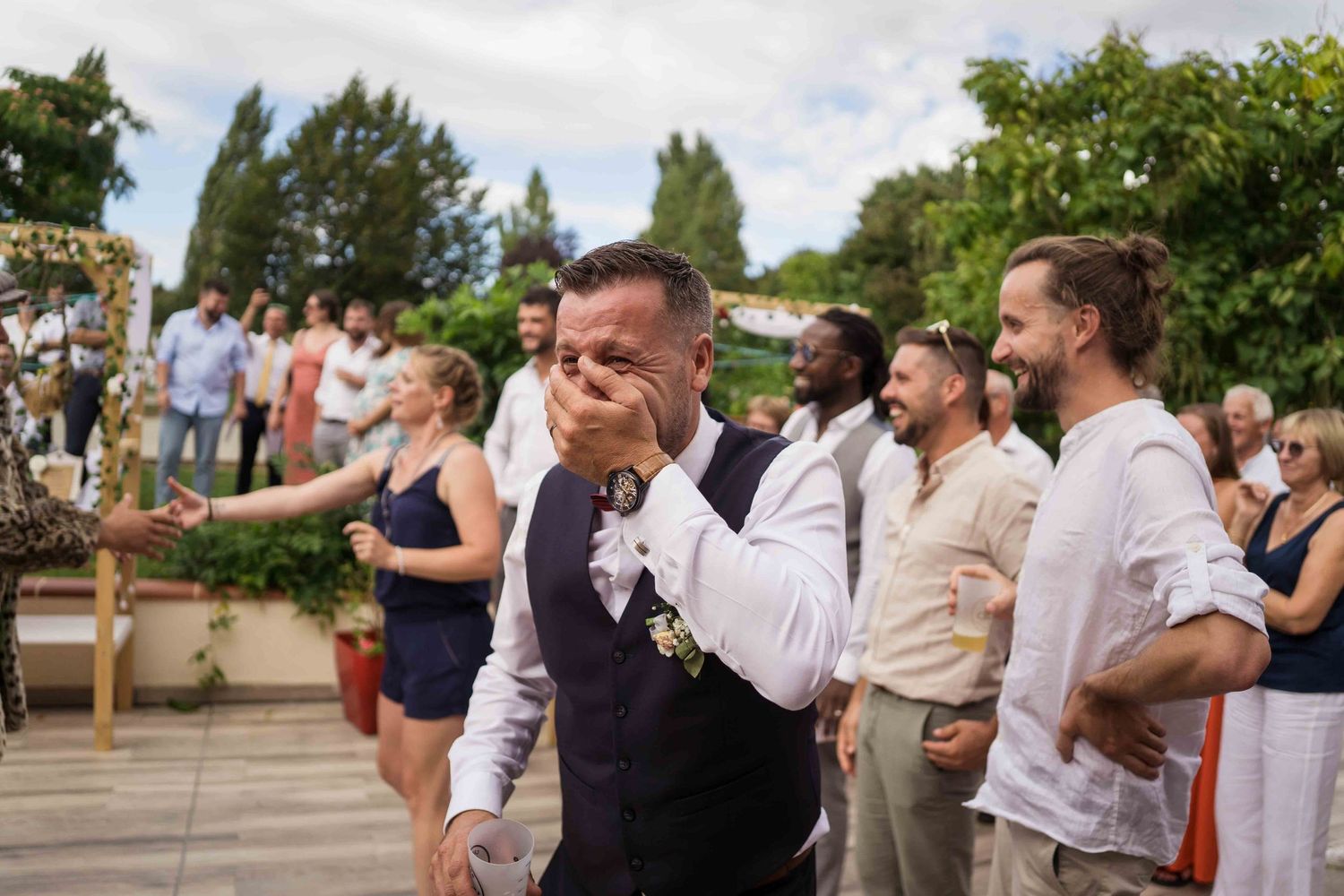 Le marié, ému, essuie ses larmes lors de la cérémonie de mariage en plein air, entouré d'invités souriants.