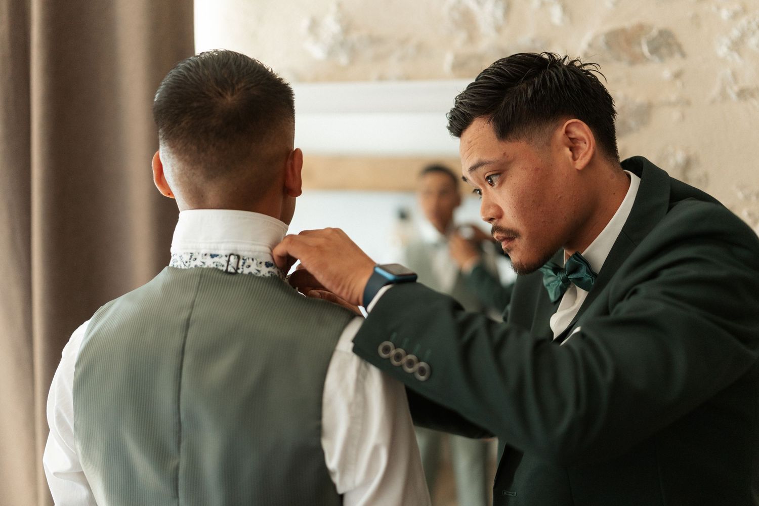 Groomsmen in green suits helping each other with collar and bow tie while getting ready for a wedding ceremony.