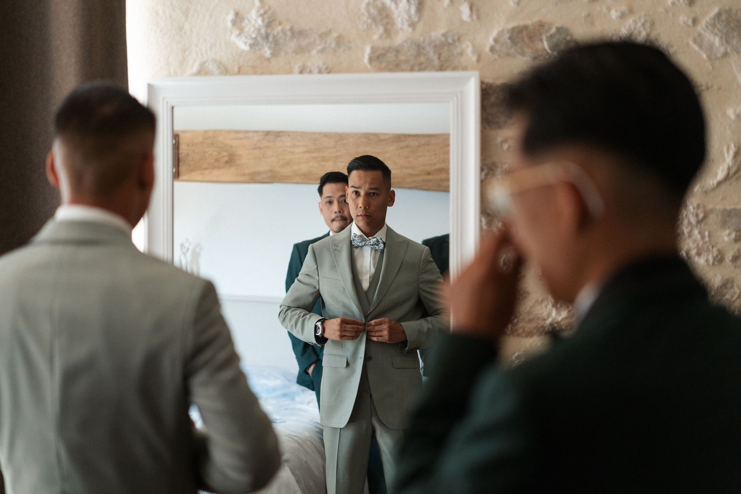 Groom in gray suit adjusts jacket while looking in mirror, with groomsmen nearby in elegant wedding preparation scene.