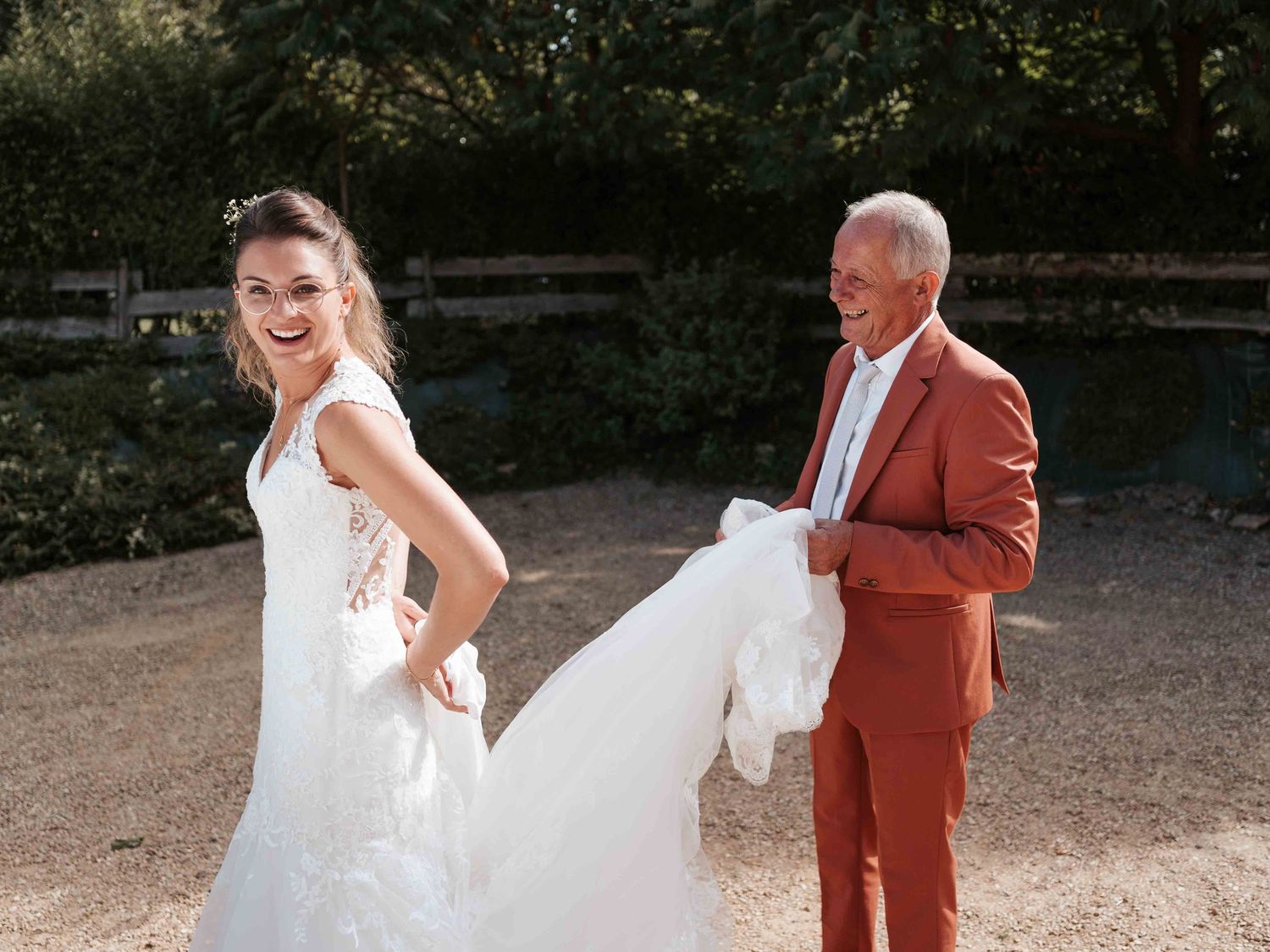 Une mariée souriante en robe de dentelle blanche rit tandis qu'un homme plus âgé en costume rouille tient sa traîne.