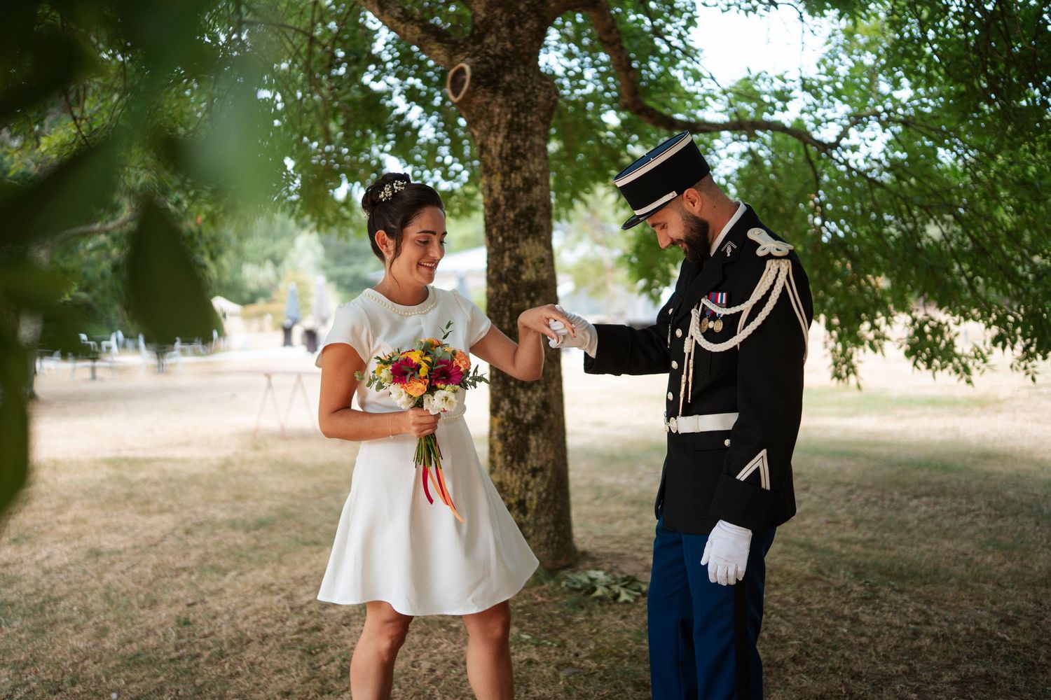Groom in military uniform kisses bride's hand outdoors under tree, bride holds colorful bouquet in white dress.
