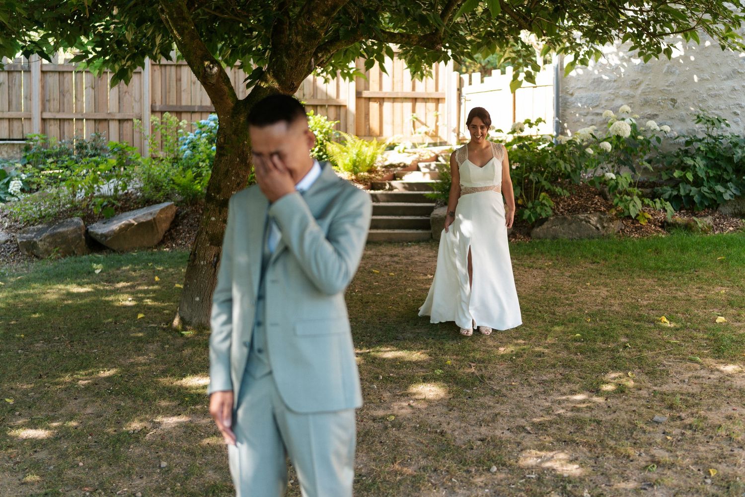 Groom in light blue suit covers his face emotionally as bride in white dress approaches for first look in garden.