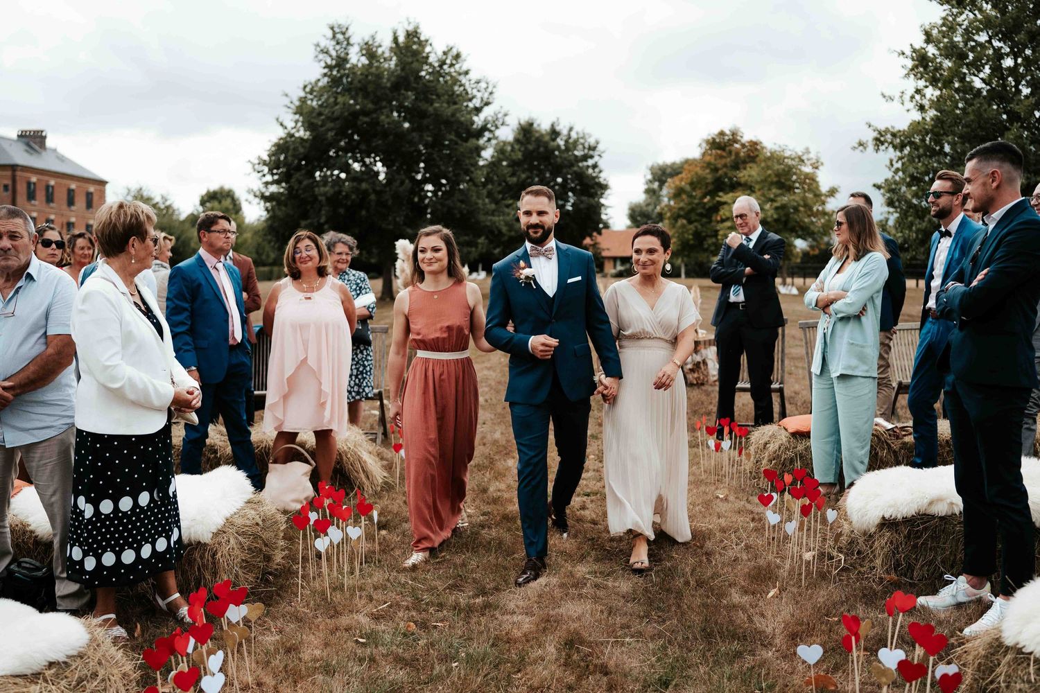 Les jeunes mariés descendent une allée extérieure rustique bordée de fleurs rouges et de bottes de foin, accompagnés de leurs invités.
